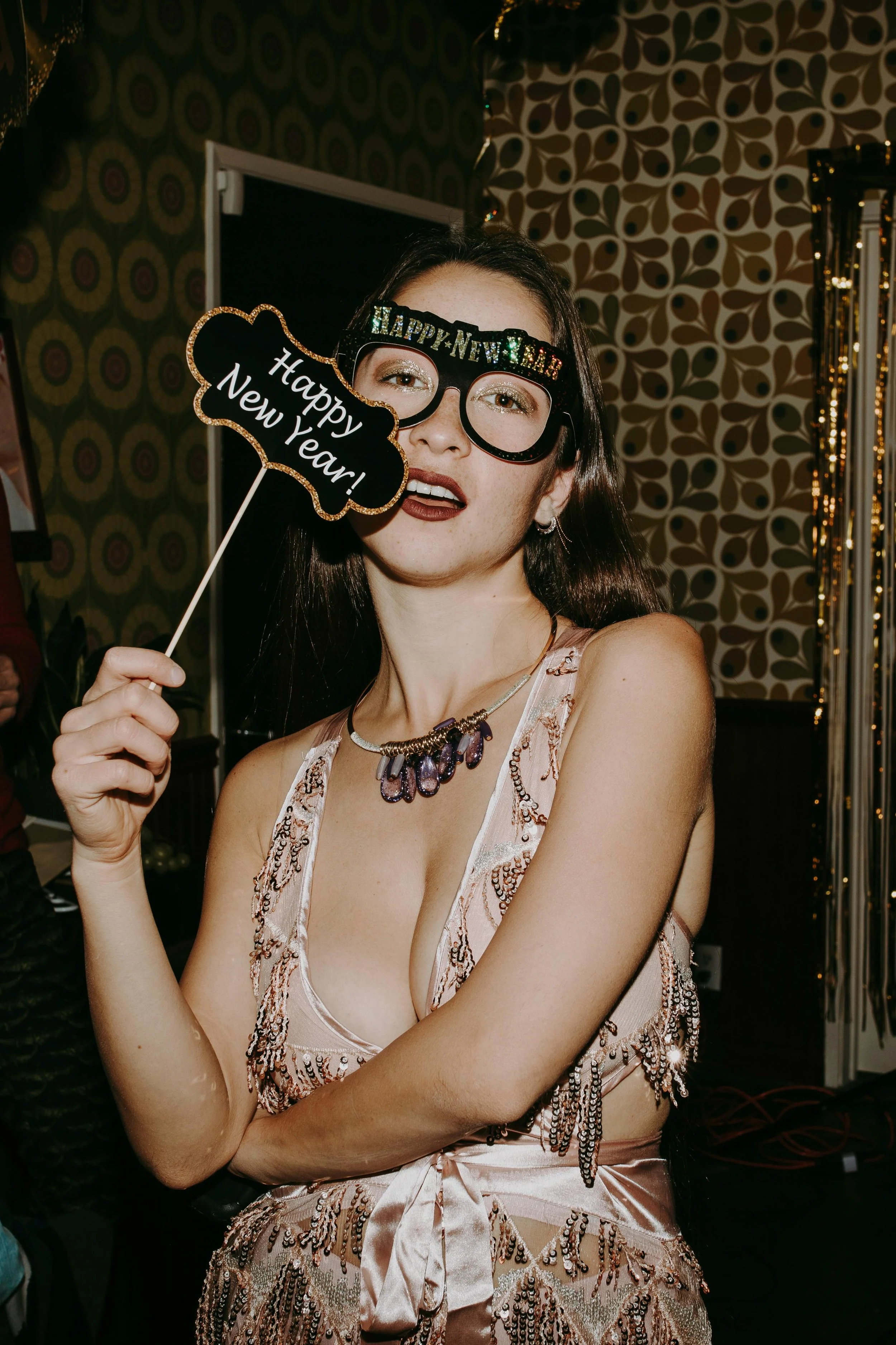 A young woman at a New Year's Eve celebration, holding a sign that says 'Happy New Year!' and wearing novelty glasses with 'HAPPY NEW YEAR' written on them. She is dressed in a pink, sequined dress with bead embellishments, and has shoulder-length dark hair, earrings, and a necklace.