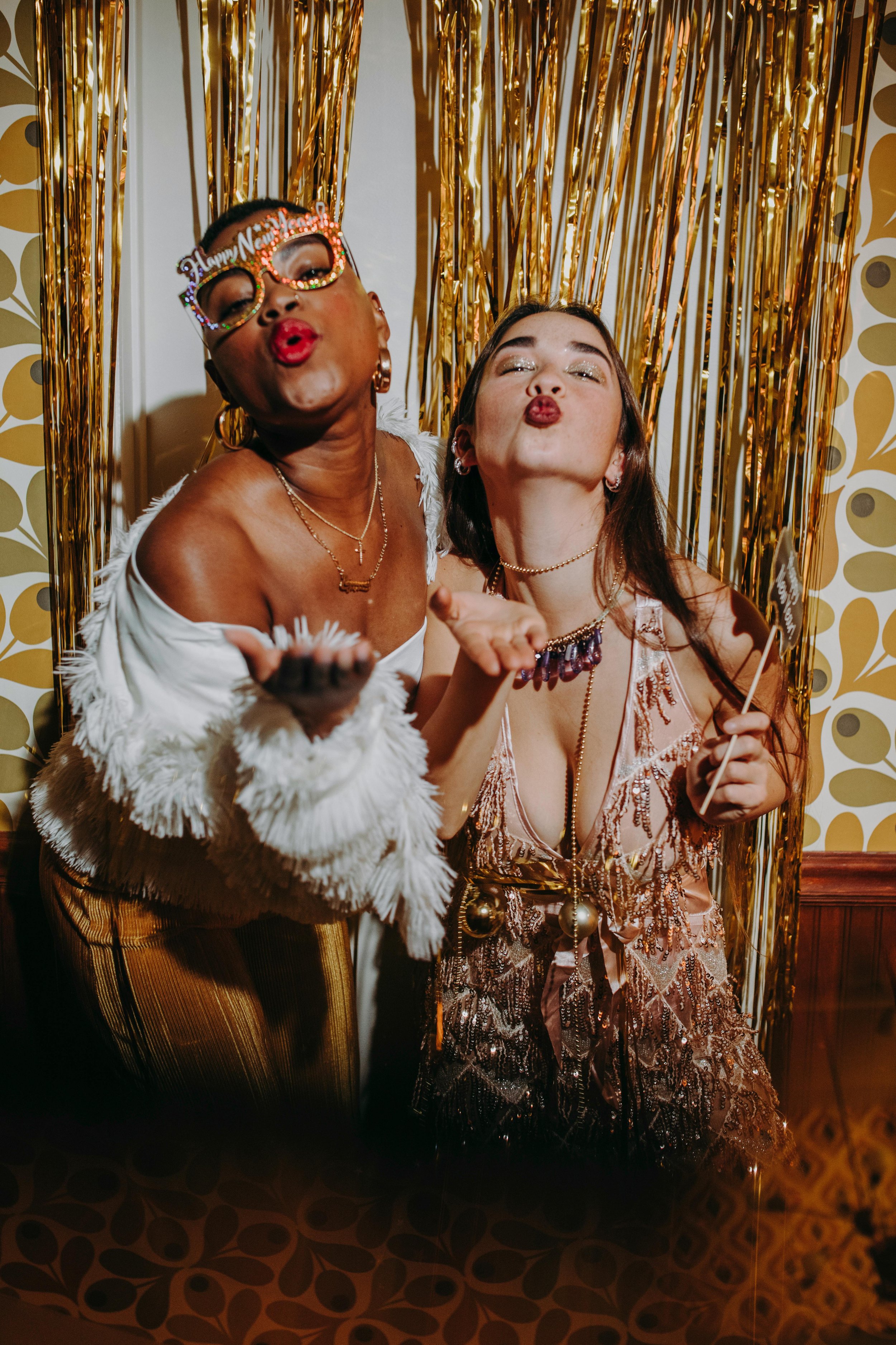 Two women blowing kisses in front of gold tinsel curtains at a New Year's celebration. One woman wears glasses with 'Happy New Year' and the other has glittery eyeshadow and jewelry.