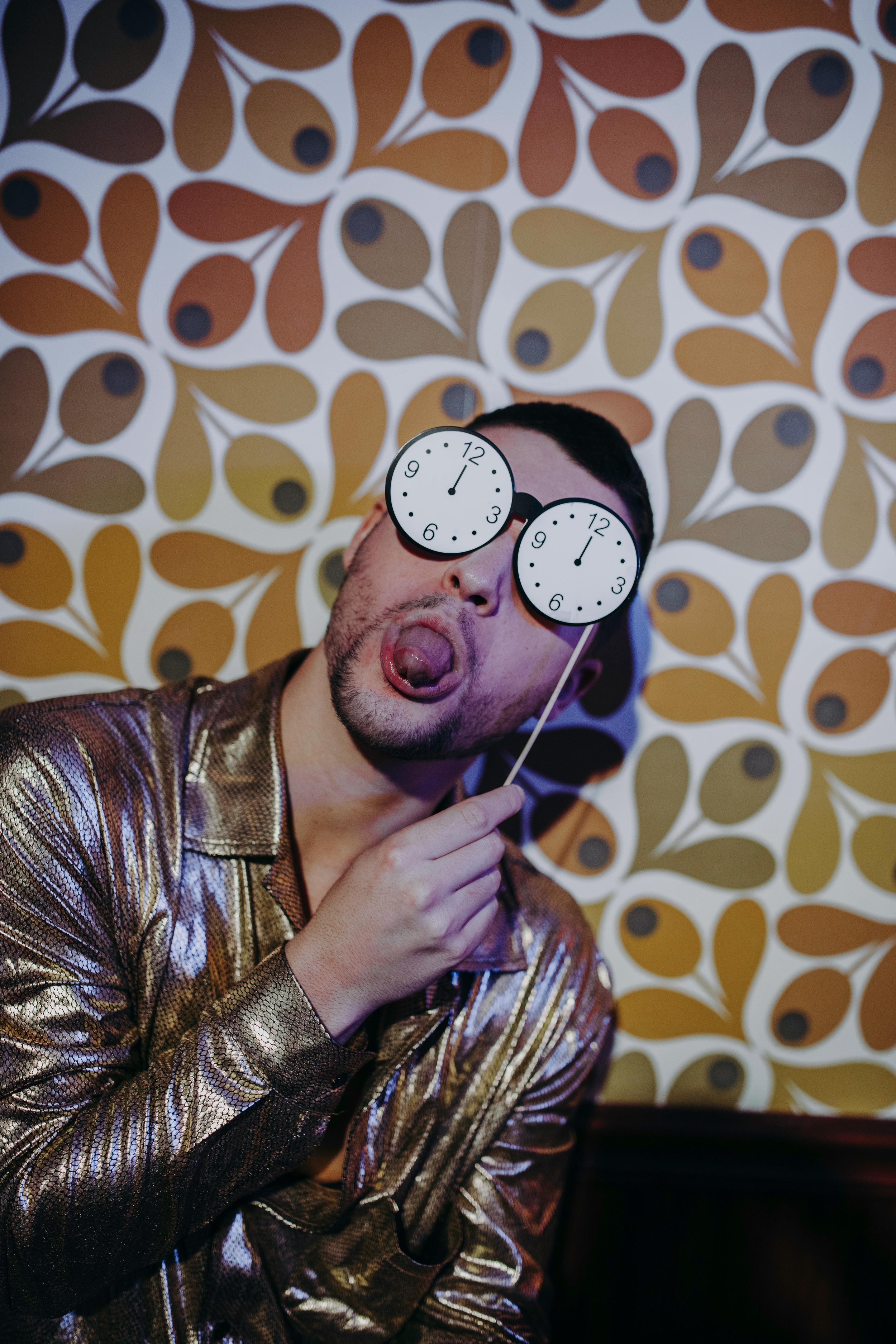 A person with short dark hair and a beard making a funny face with tongue out, wearing novelty sunglasses with clock faces, in front of a retro patterned wallpaper with brown, beige, and white abstract shapes.