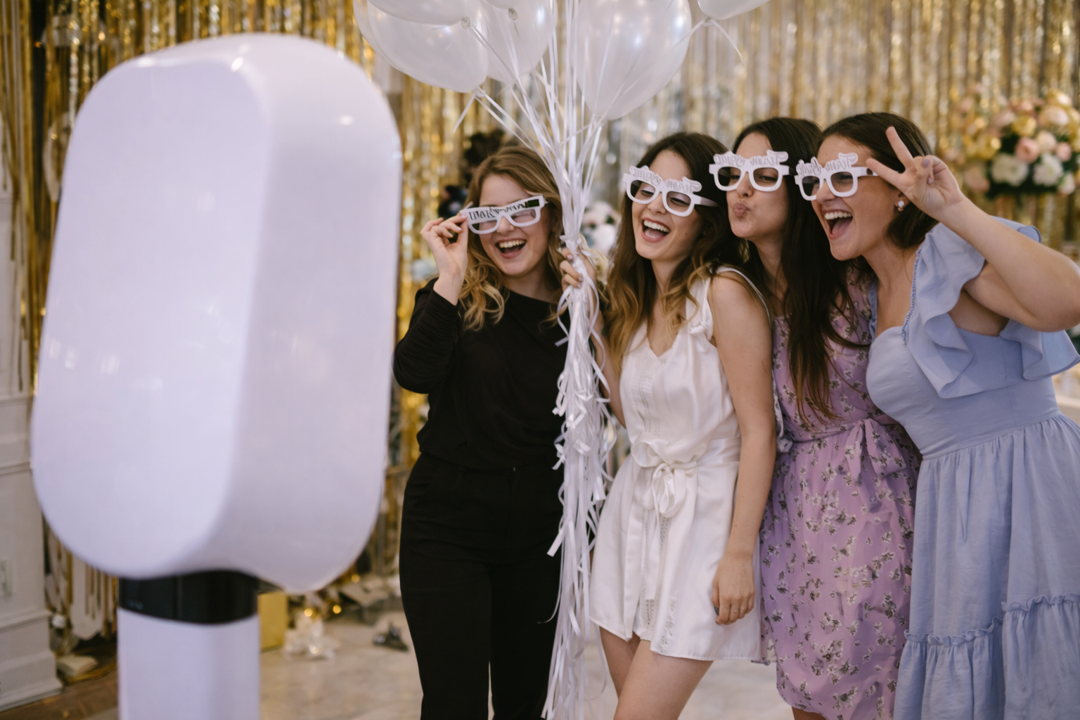 Four women celebrating at a party, wearing white and pink novelty glasses, smiling and posing with a bunch of white balloons against a gold backdrop.