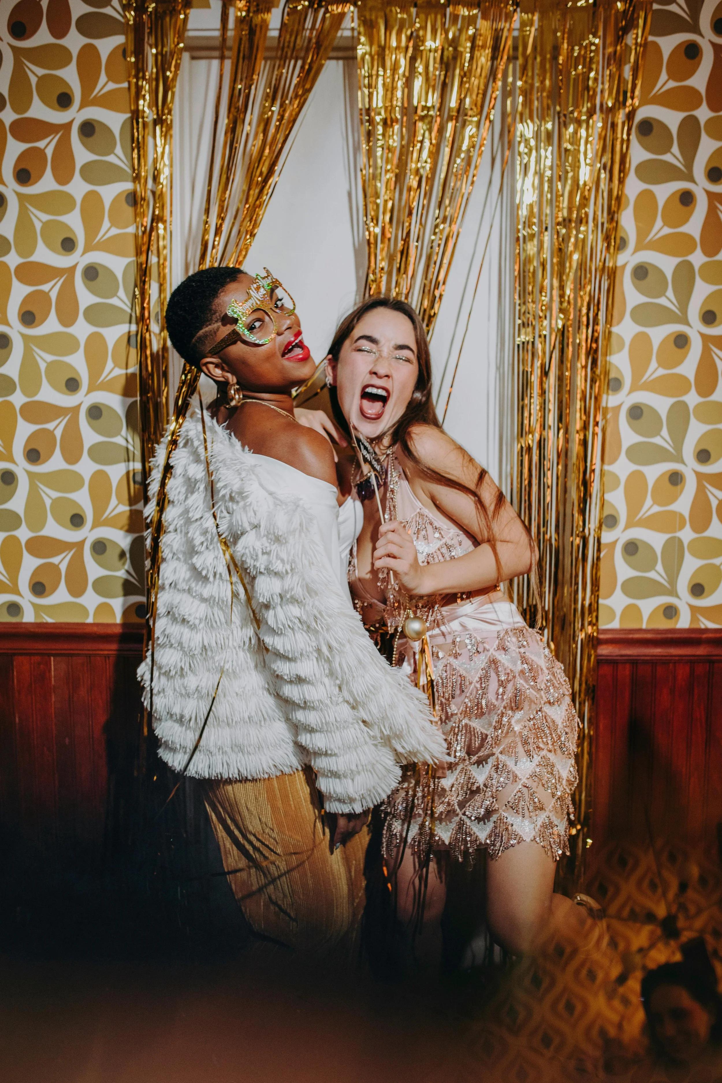 Two women dressed in glamorous attire dancing or celebrating at a party with gold and black decorations in the background.