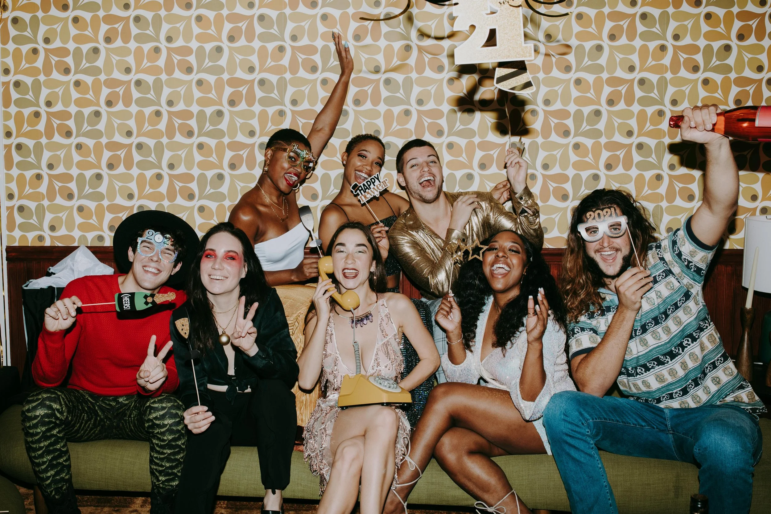 A group of friends celebrating New Year's Eve at a party, wearing party accessories, and holding drinks and party props in front of a patterned wallpaper.