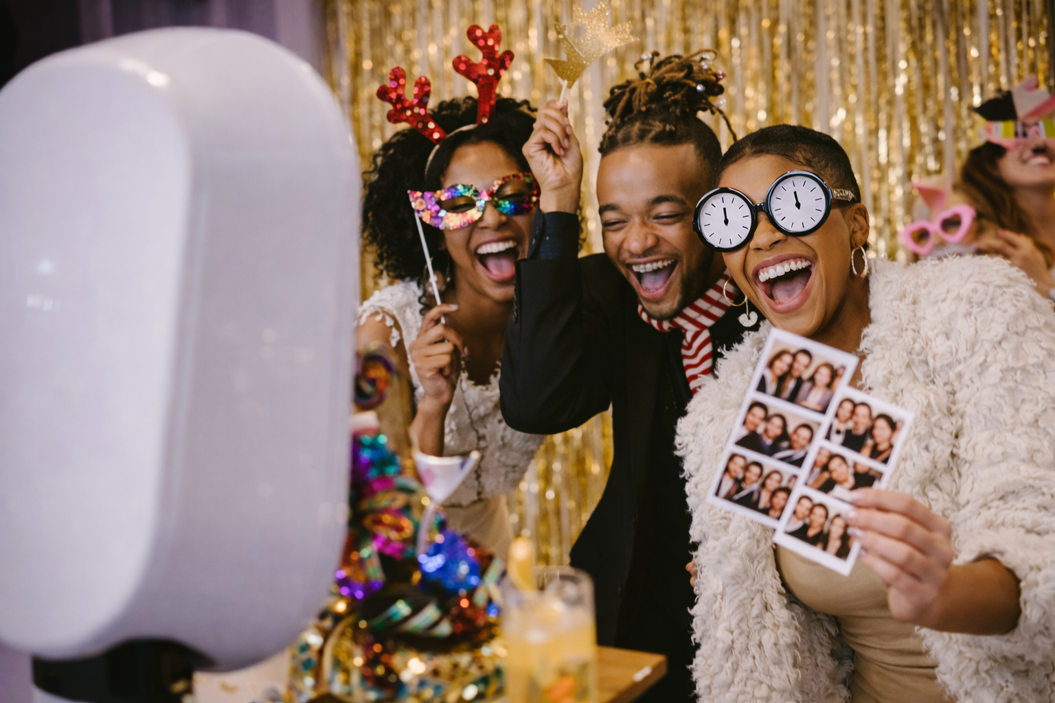 A group of four friends celebrating New Year's Eve at a party, wearing fun costumes and photobooth props, including clocks and glasses, smiling and taking selfies in front of a gold sequin backdrop.