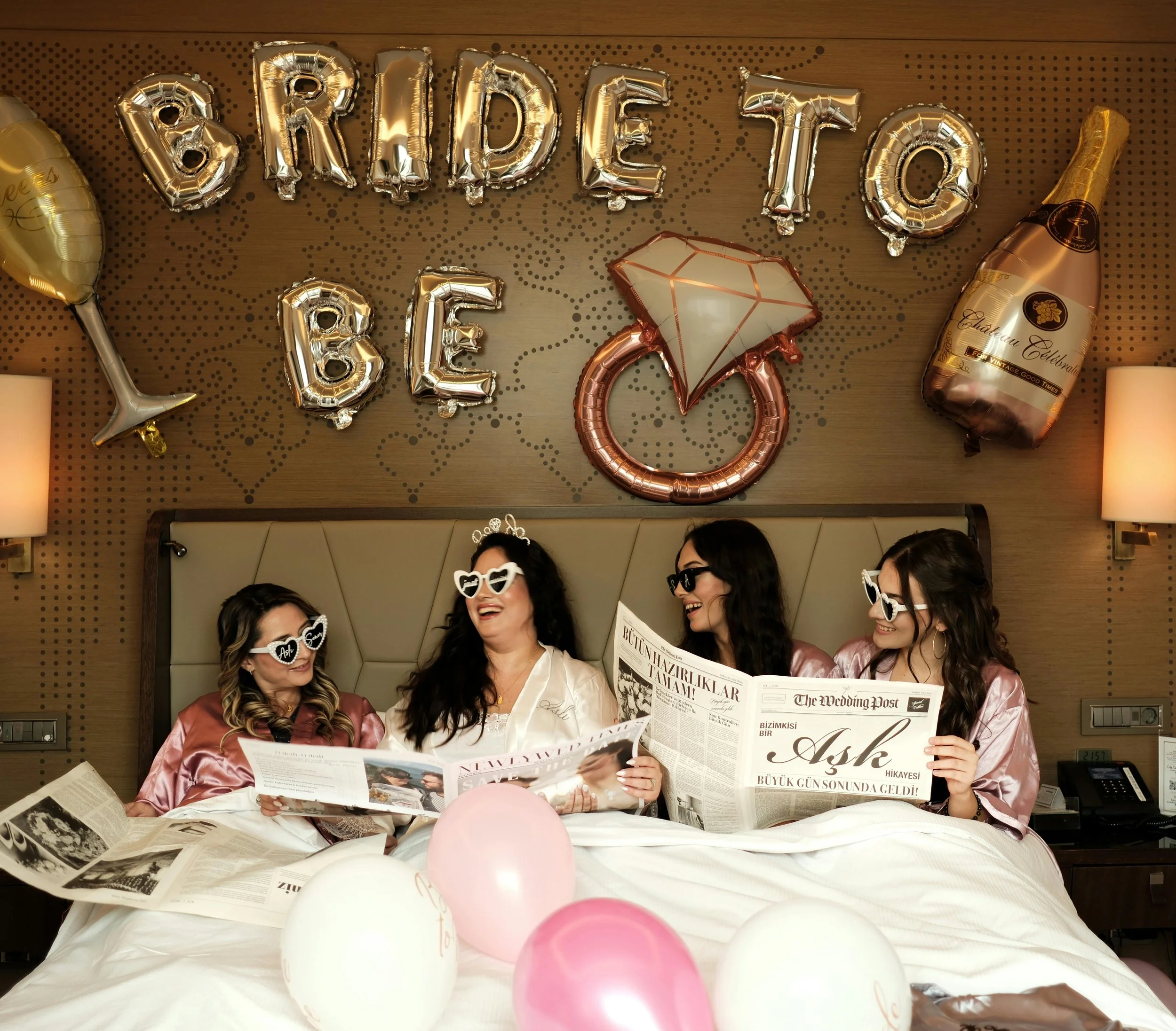 Women sitting on a bed with balloons, reading newspapers, with balloons spelling 'BRIDE TO BE' on the wall and celebratory decorations.