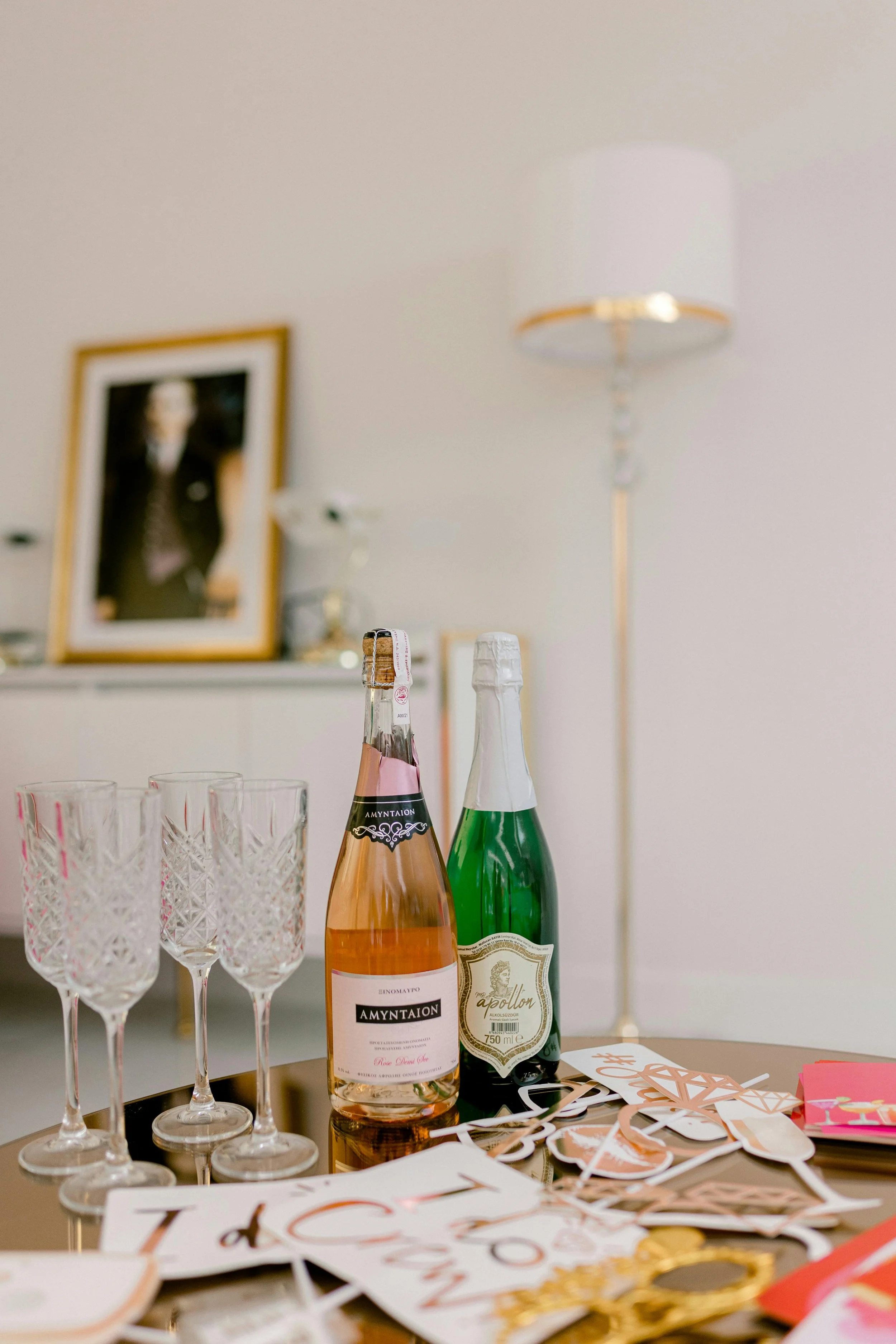 A table containing two bottles of sparkling wine, three empty crystal wine glasses, and various party decorations including paper cutouts and confetti, with a blurry framed photograph and a lamp in the background.