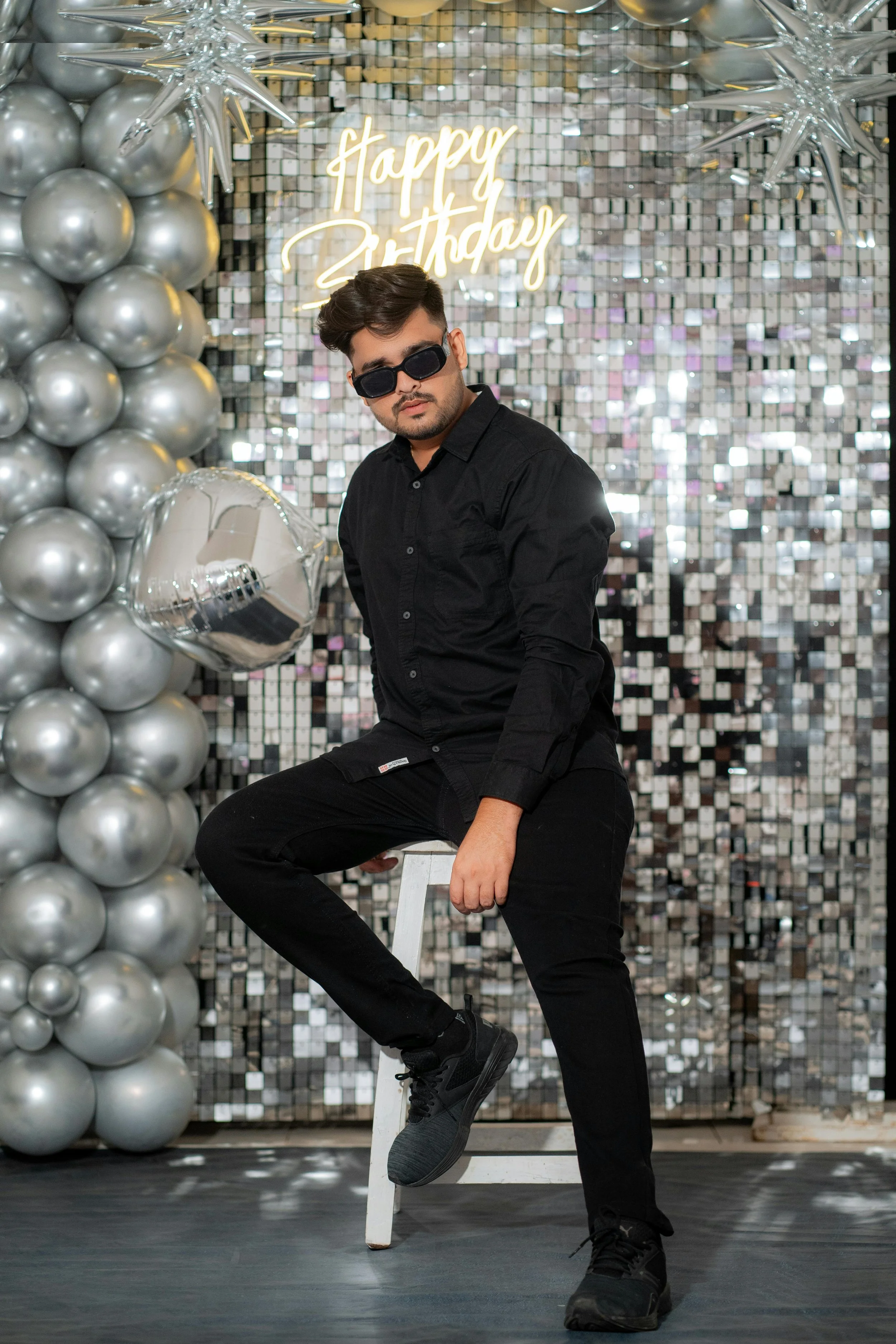 A young man dressed in all black, wearing sunglasses, sitting on a white stool against a reflective silver tile backdrop with a neon sign that says "happy birthday." To his left are silver balloons arranged in a cluster, including a star-shaped ballo