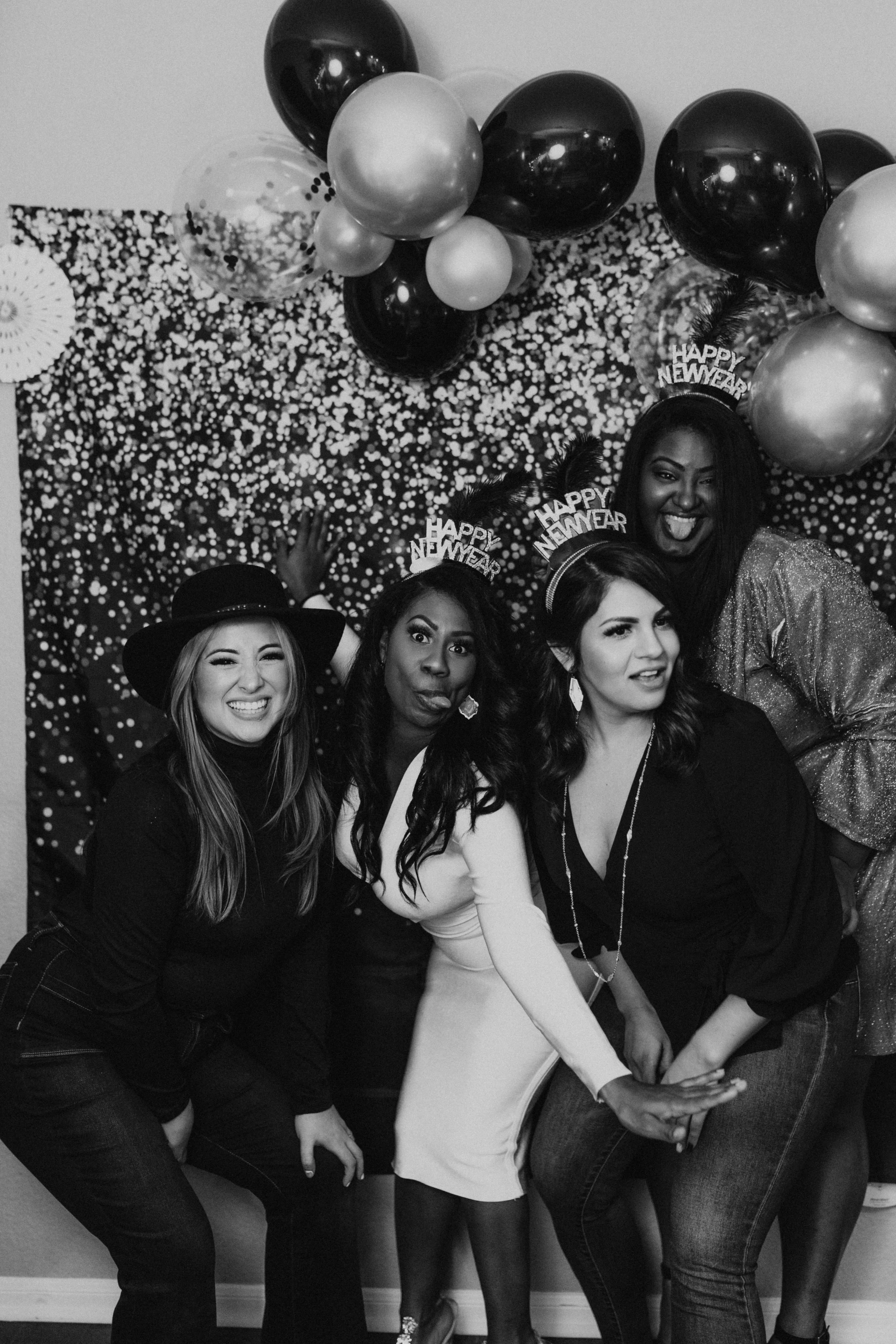Group of women at a New Year's celebration, with balloons and a festive backdrop. One woman is wearing a 'Happy New Year' headband and sticking her tongue out.