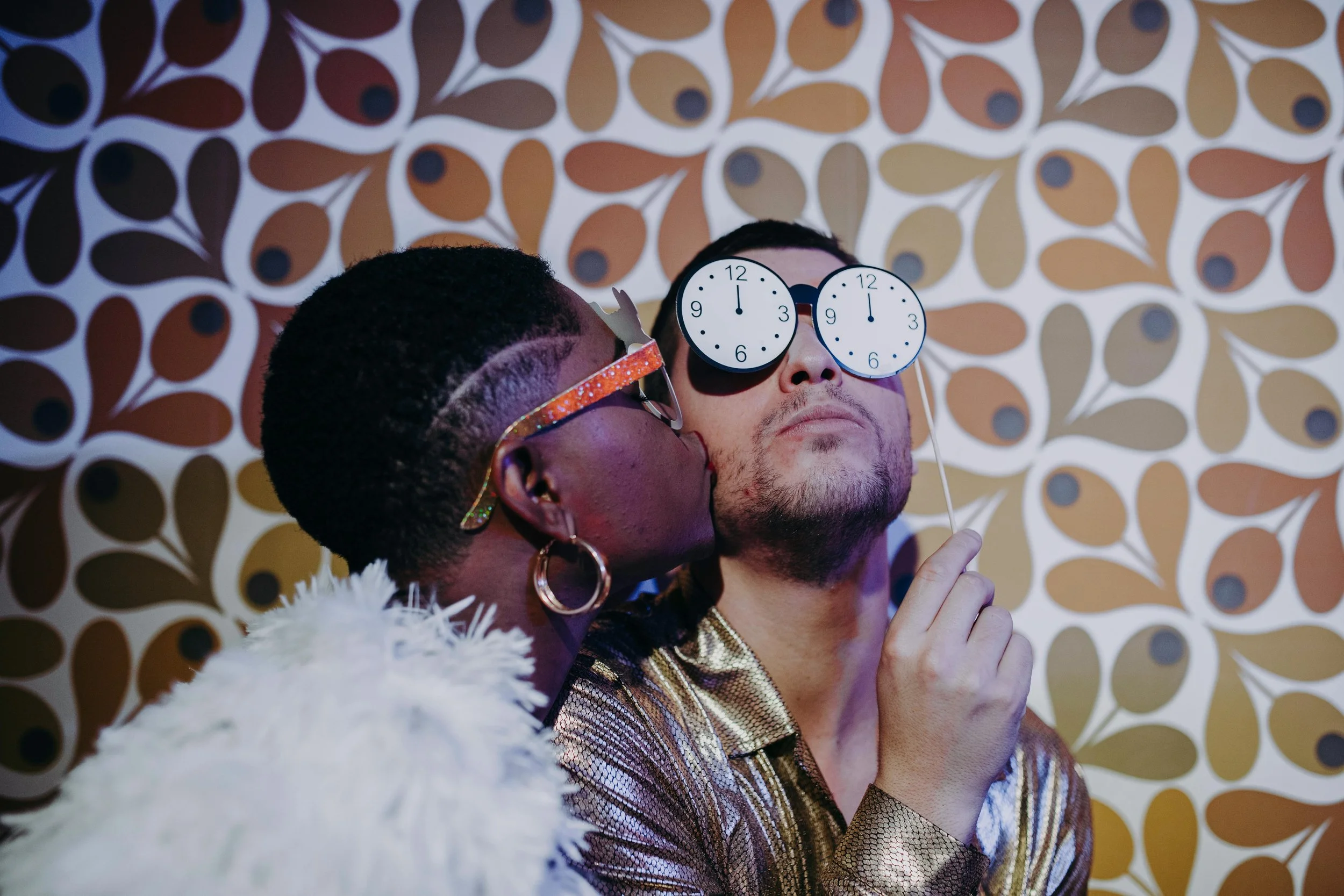 Two men taking a selfie, one is wearing novelty glasses with clock faces, and the other wearing glasses with a glittery orange frame. They are in front of a retro wallpaper pattern with teardrop shapes in brown, beige, and blue. The man in the foreground has earrings and a gold and black checkered shirt, and the other has short curly hair and is wearing a white furry coat.