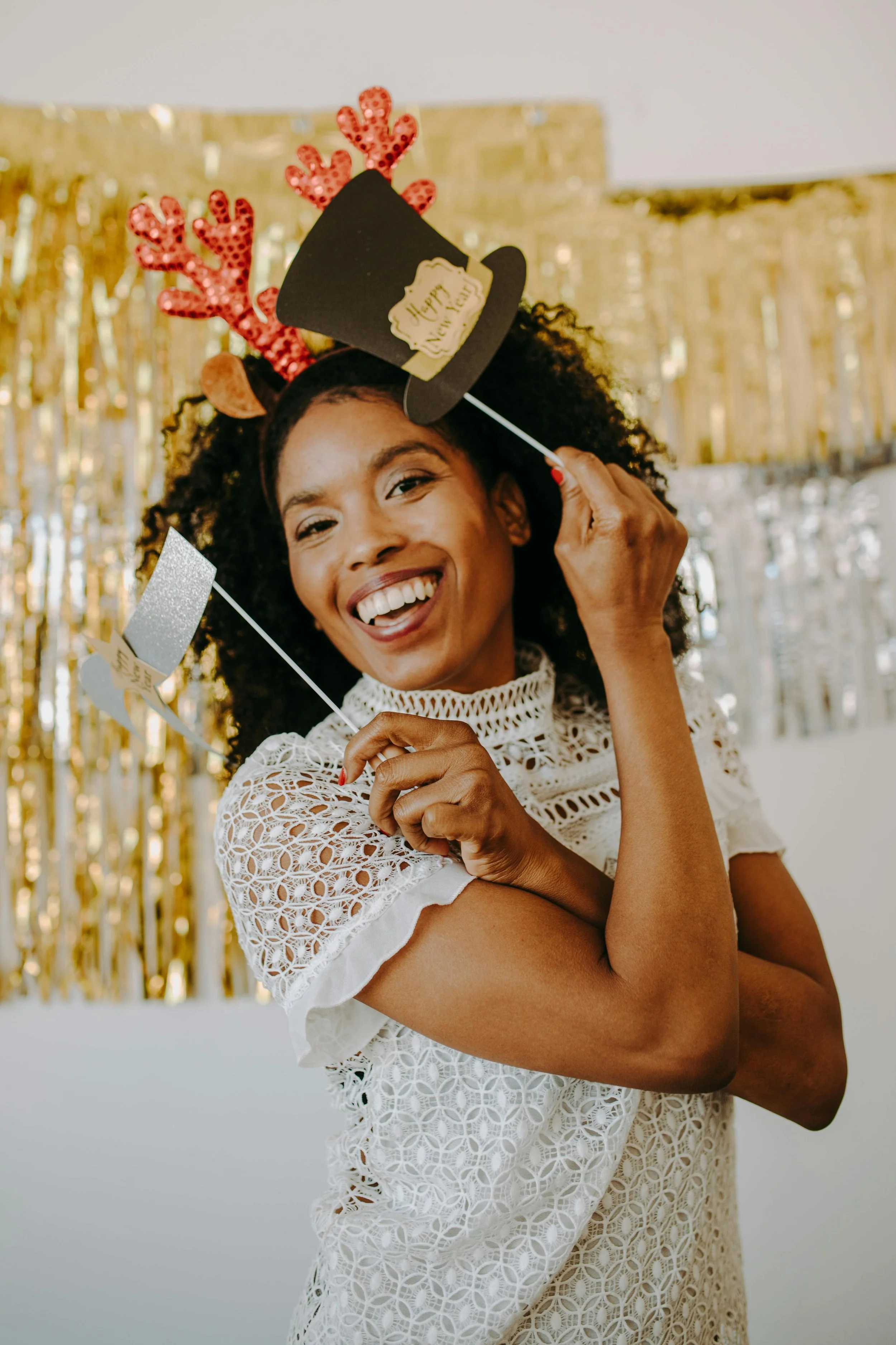 A woman with curly hair, smiling, wearing a white lace dress, holding a party photo booth prop that says 'Happy New Year,' and wearing reindeer antlers with red decorations, standing in front of a gold foil curtain.