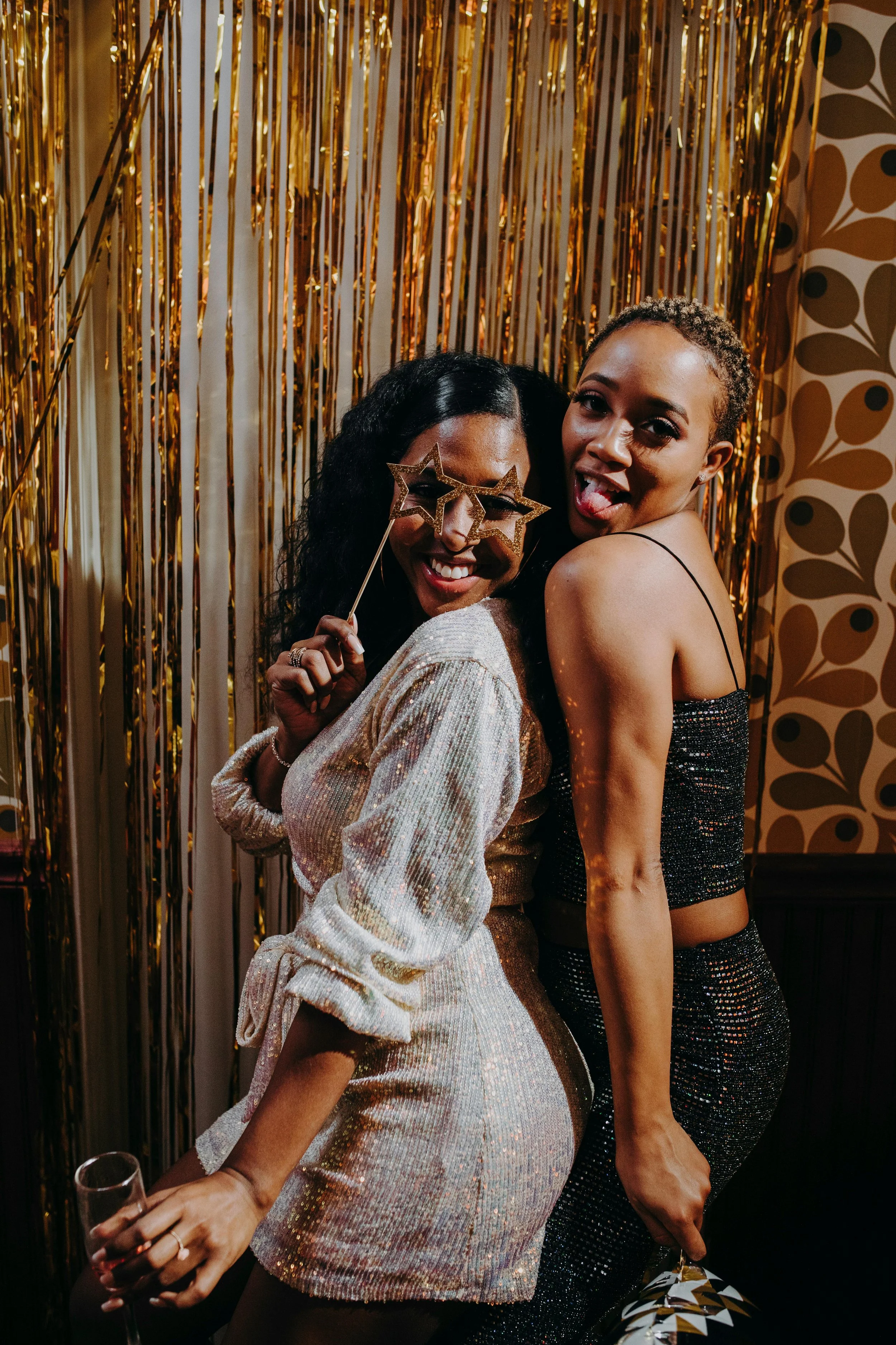 Two women at a party posing in front of a gold and black streamers backdrop. One woman is wearing star-shaped glasses, holding a glass of wine, and smiling; the other is sticking out her tongue, wearing a black, sparkly dress, and has her arm around 