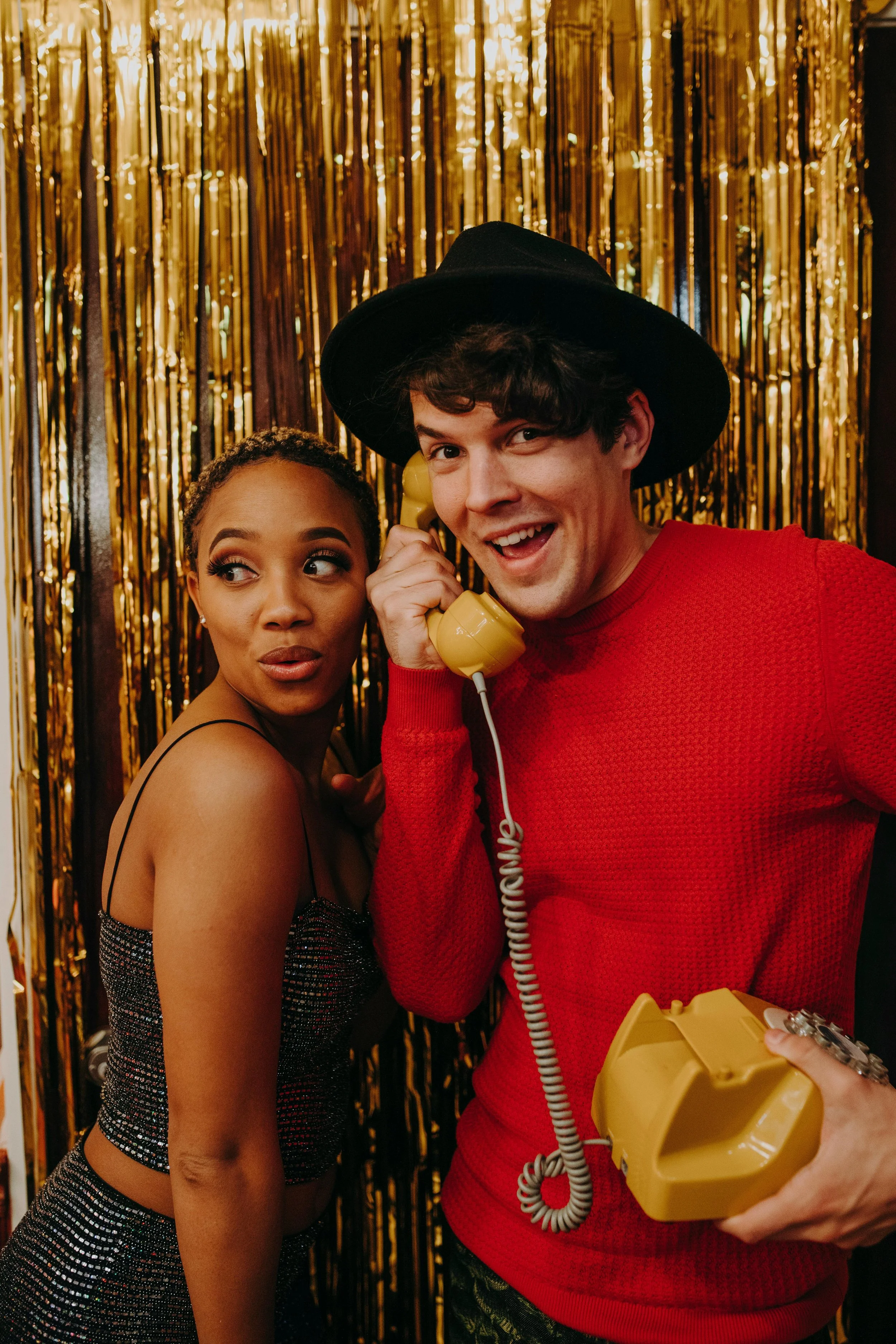 A woman with makeup and a sleeveless black dress standing next to a man wearing a red sweater, black hat, and holding a yellow rotary phone, against a gold tinsel backdrop.