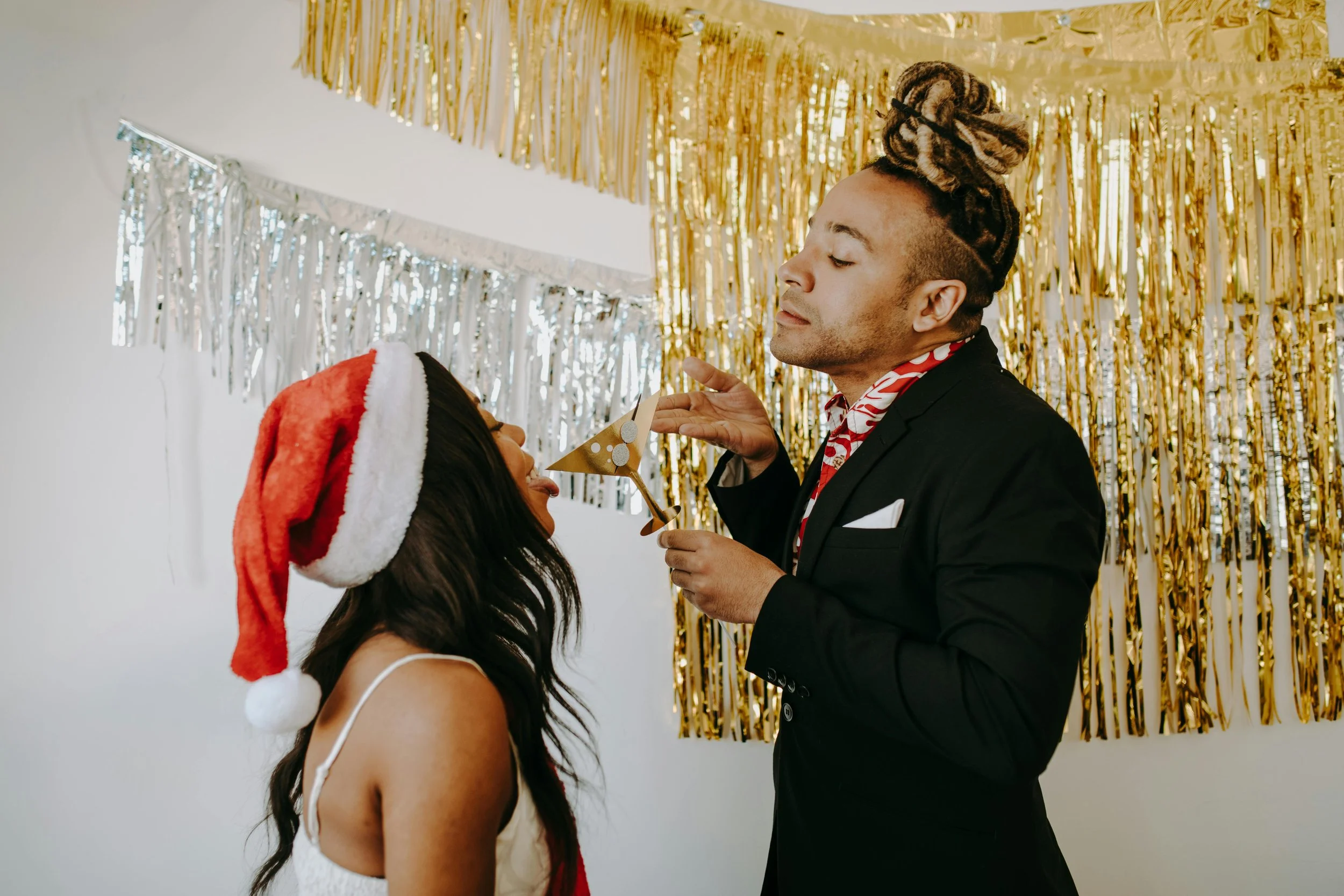 A man and woman face each other during a festive celebration, with the man holding a party horn in front of the woman. The woman wears a red Santa hat and a white dress, and the man has dreadlocks styled in an updo, dressed in a black suit with a red and white scarf. The background features shiny gold and silver streamer decorations.