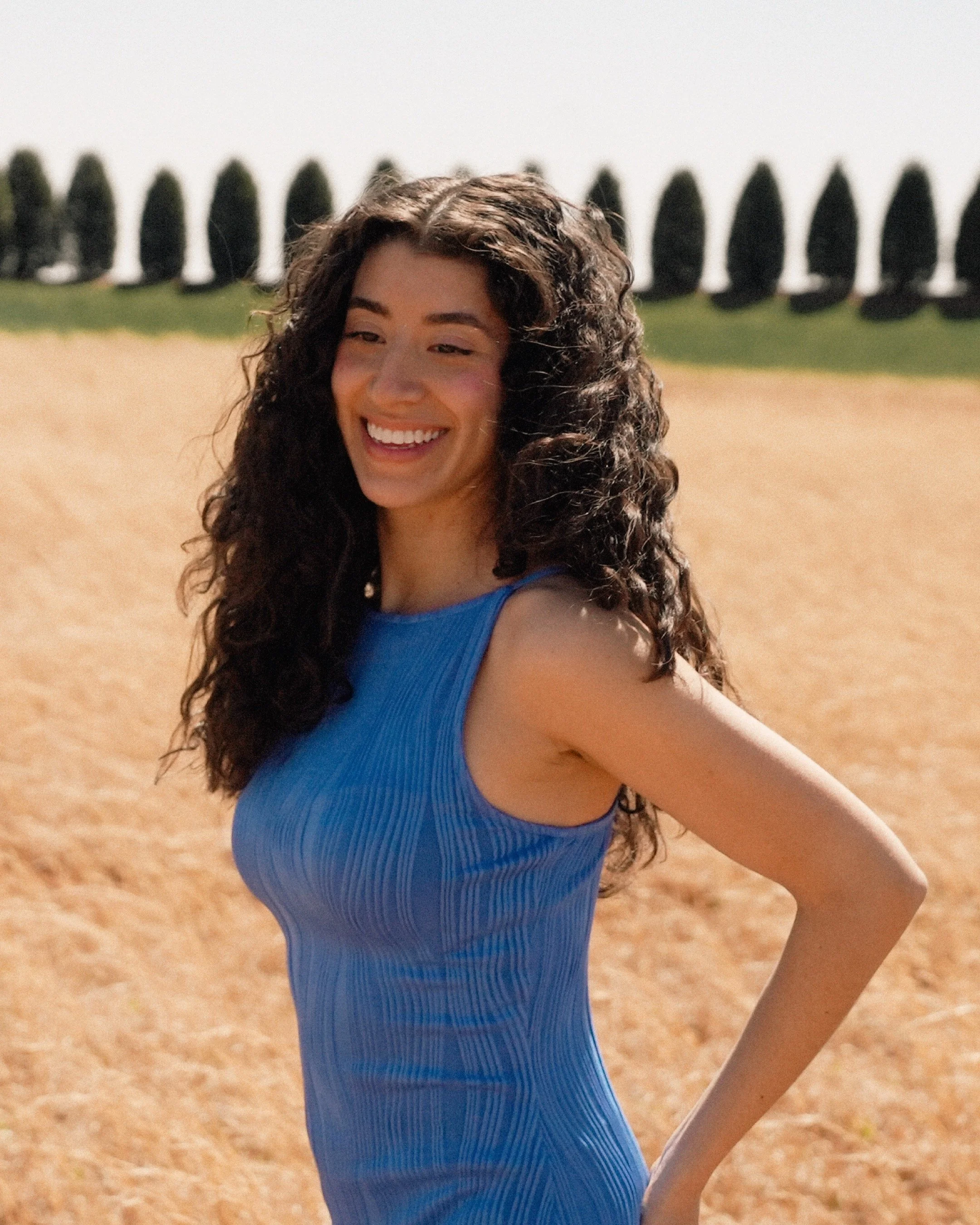 Woman with long curly dark hair wearing a sleeveless blue dress, smiling outdoors in a field with a row of tall trees in the background.