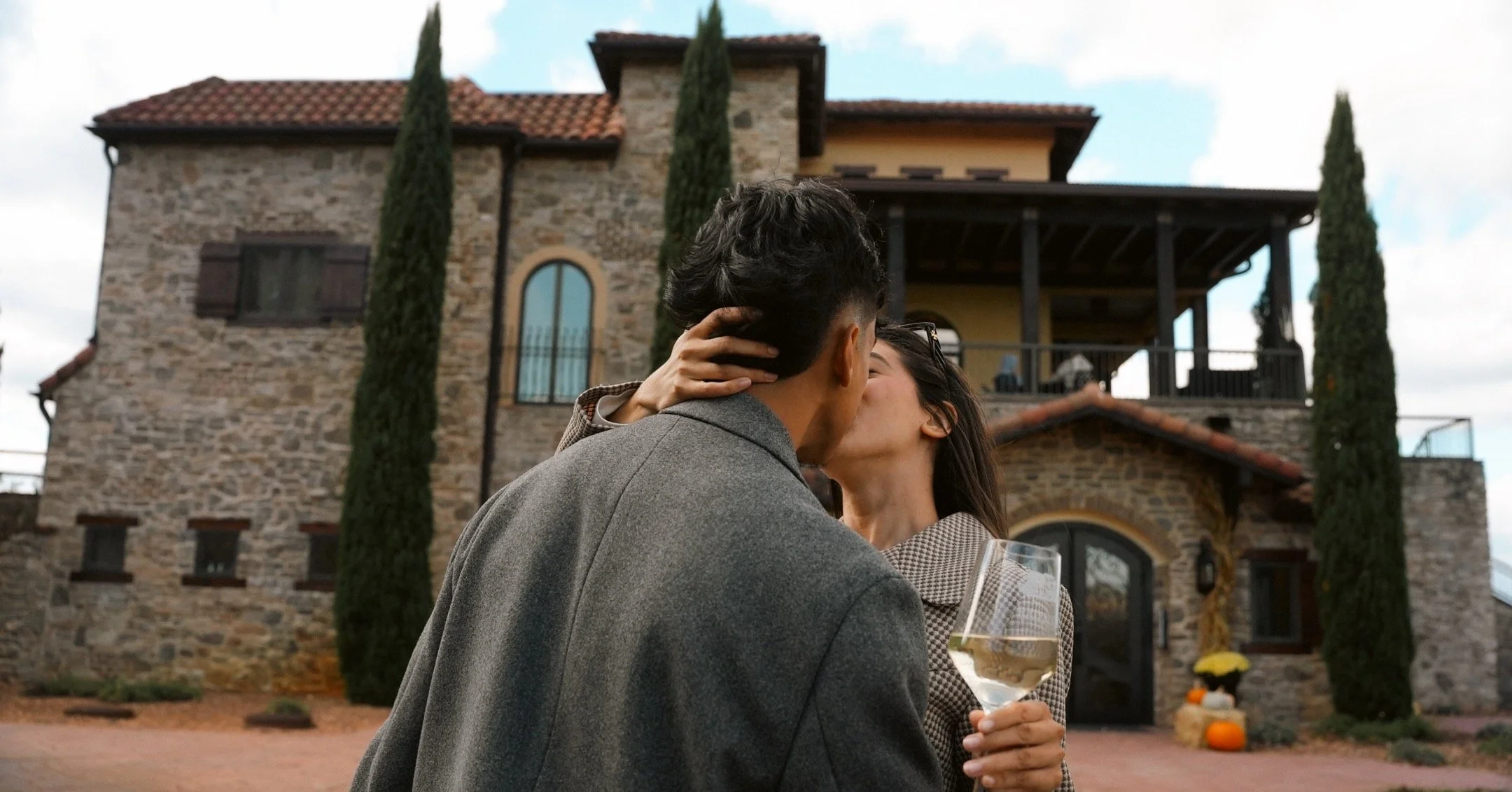 Couple kissing outdoors in front of a large stone house with tall trees and pumpkins, holding a glass of white wine.