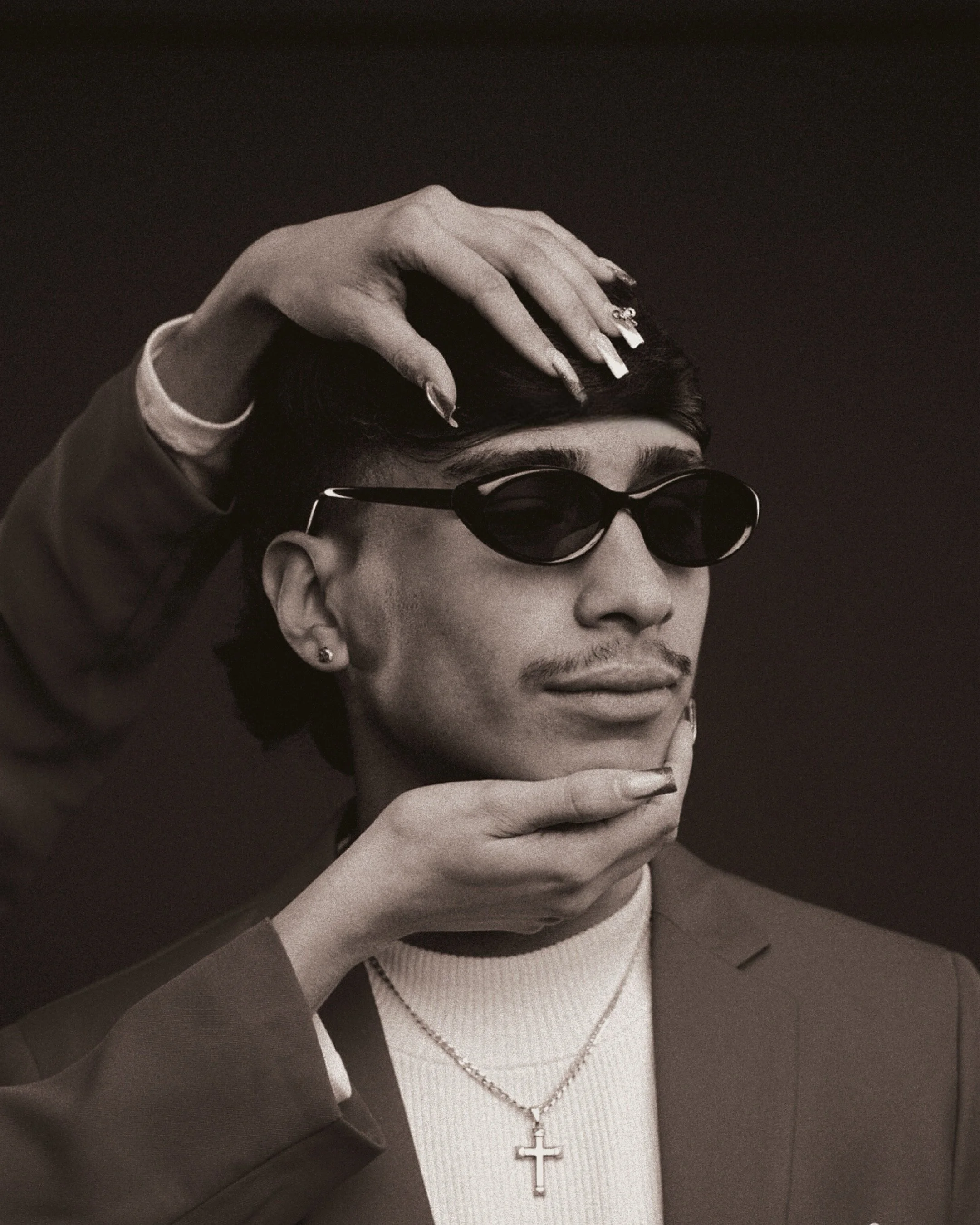 A black and white portrait of a young man with short dark hair, wearing sunglasses, a ring, earrings, a chain with a cross pendant, a turtleneck sweater, and a blazer. Someone's hands are adjusting his hair and chin.