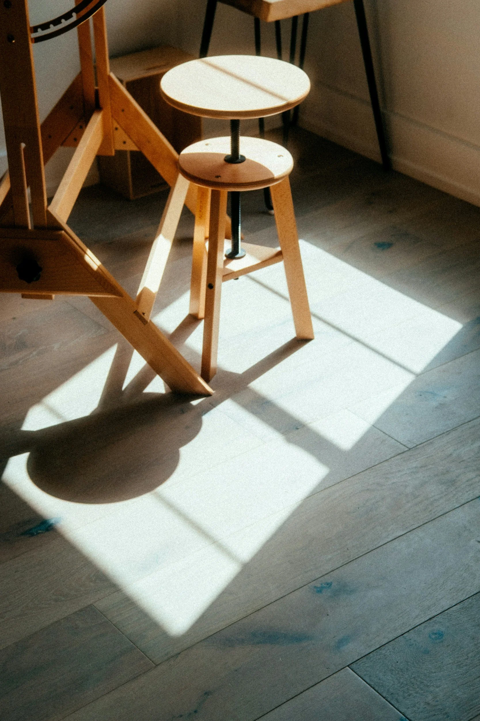 A wooden stool with a circular top and a screw in the middle, positioned on a light-colored wooden floor with sunlight casting shadows. Part of a wooden structure and a black chair are also visible.