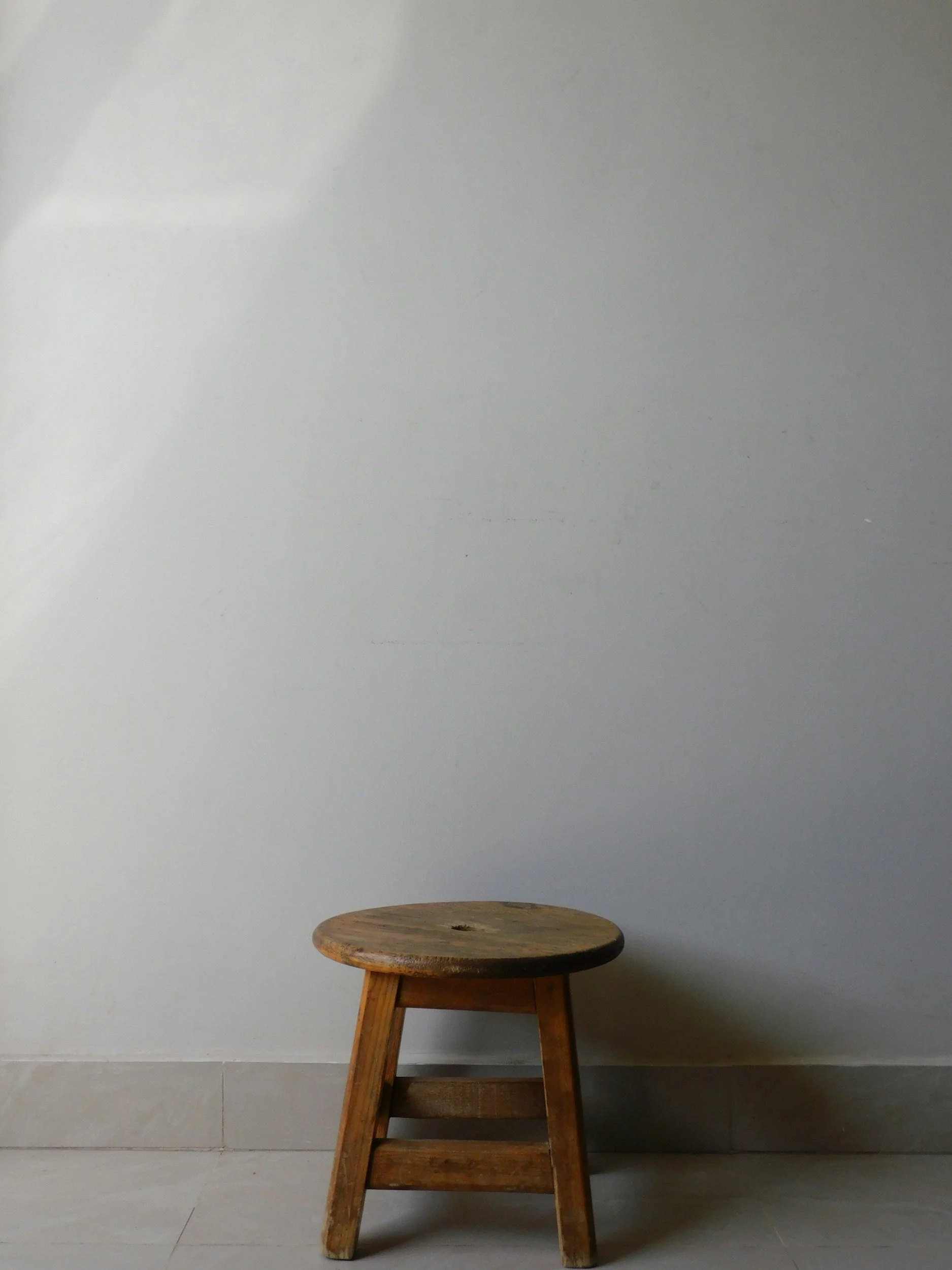 A wooden stool with four legs and a round top against a plain wall.