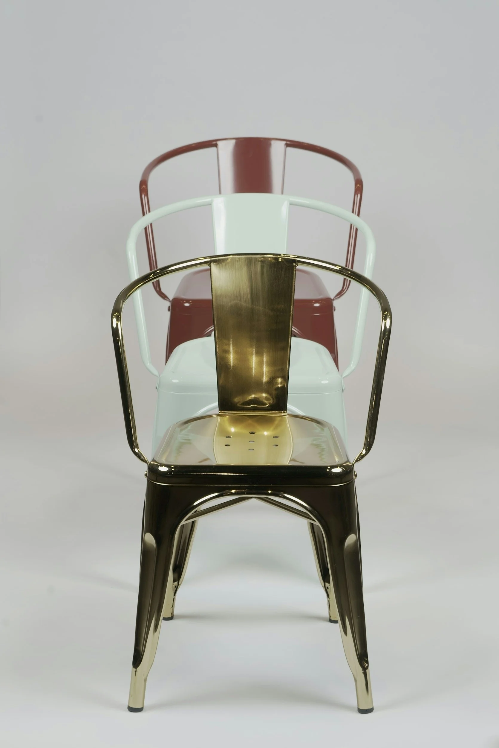 A collection of four modern metal chairs stacked one behind the other, each with a different finish: gold, white, pink, and dark brown, arranged against a plain light gray background.