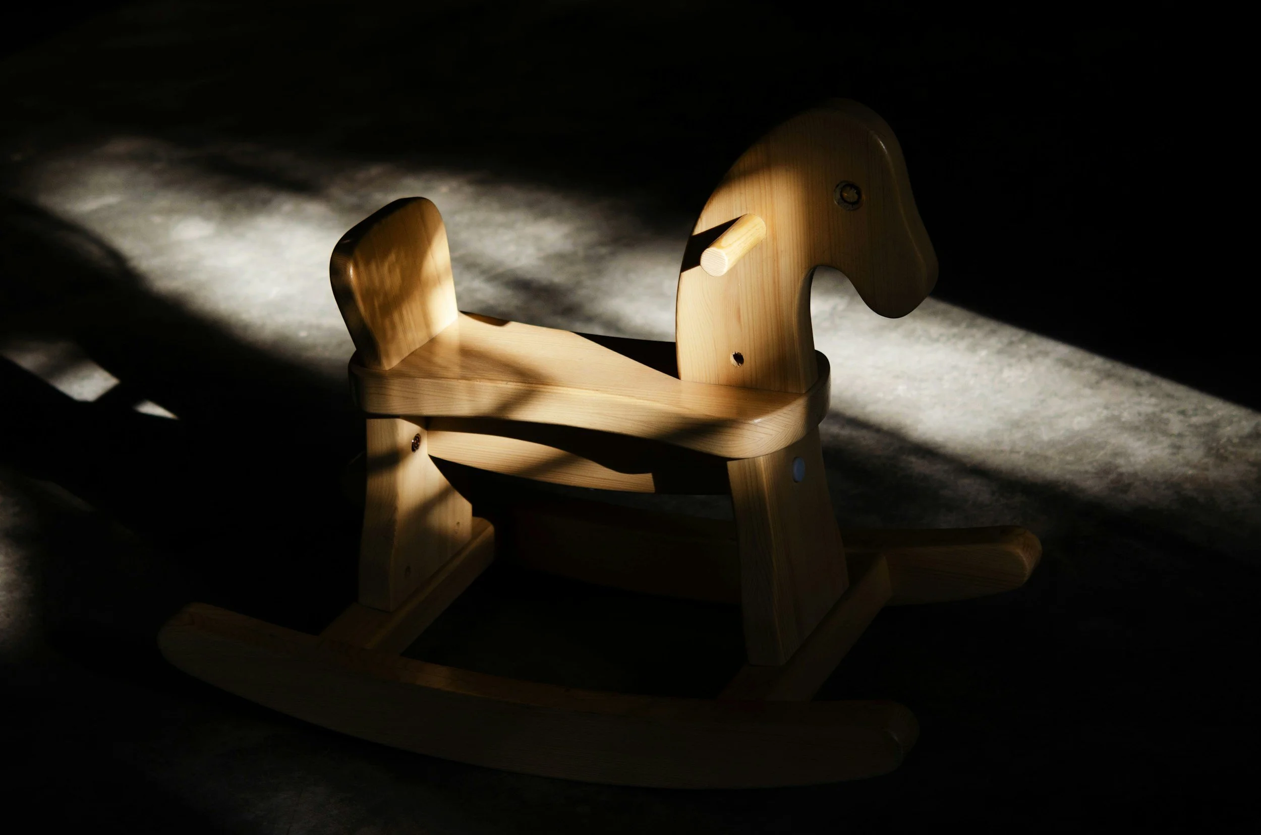 A wooden rocking horse in a dark room with light casting shadows on the floor.