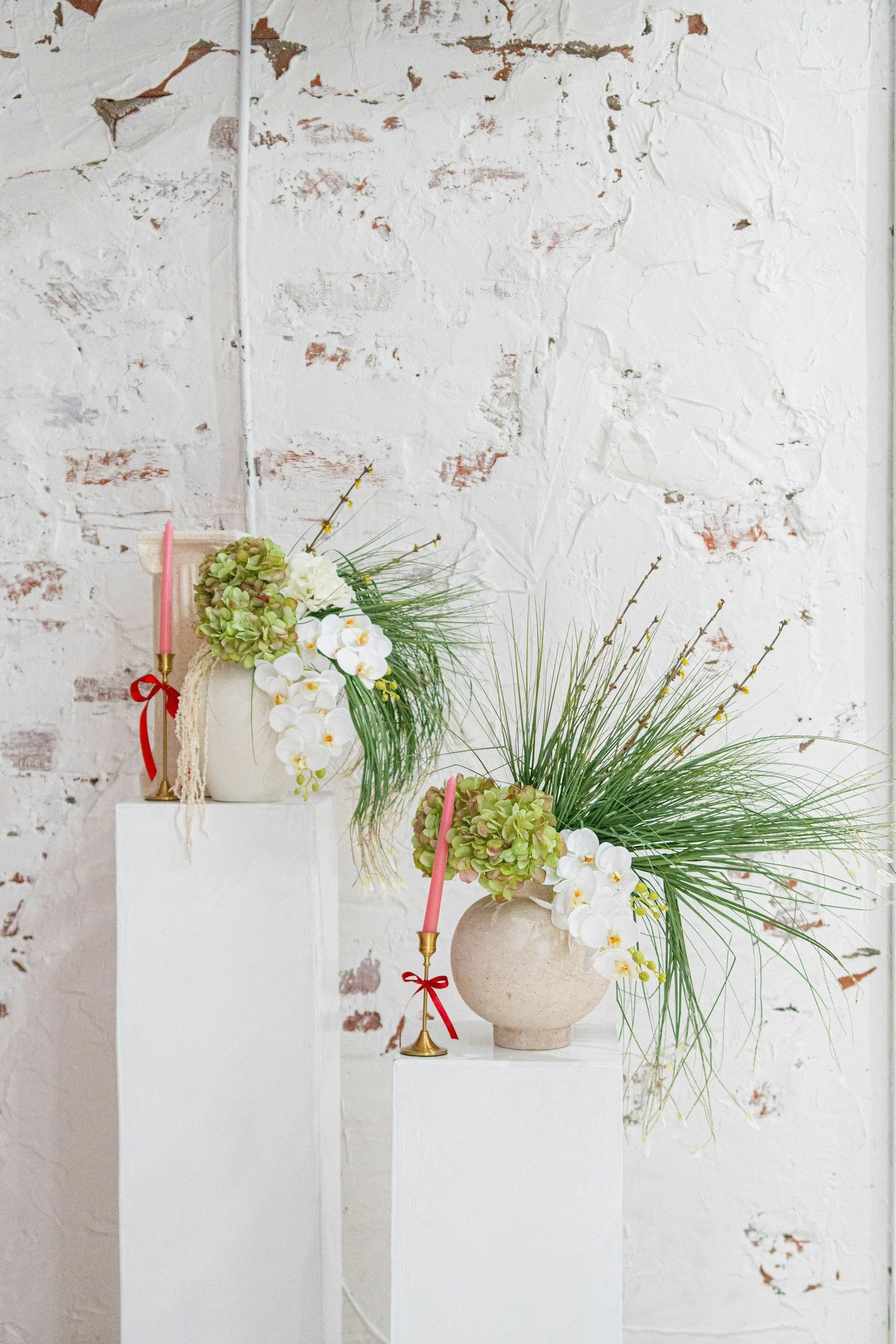Three white pedestals with floral arrangements in neutral-colored and white vases, featuring white orchids and green foliage, in front of a distressed white brick wall.