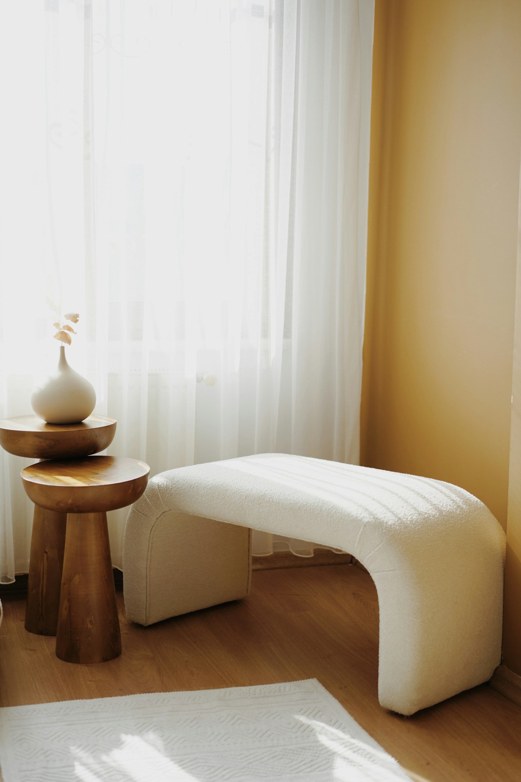 A cozy bedroom corner with sheer curtains letting in sunlight, a round wooden side table with a white vase holding dried leaves, an off-white textured chaise lounge, and a white rug on a wooden floor.