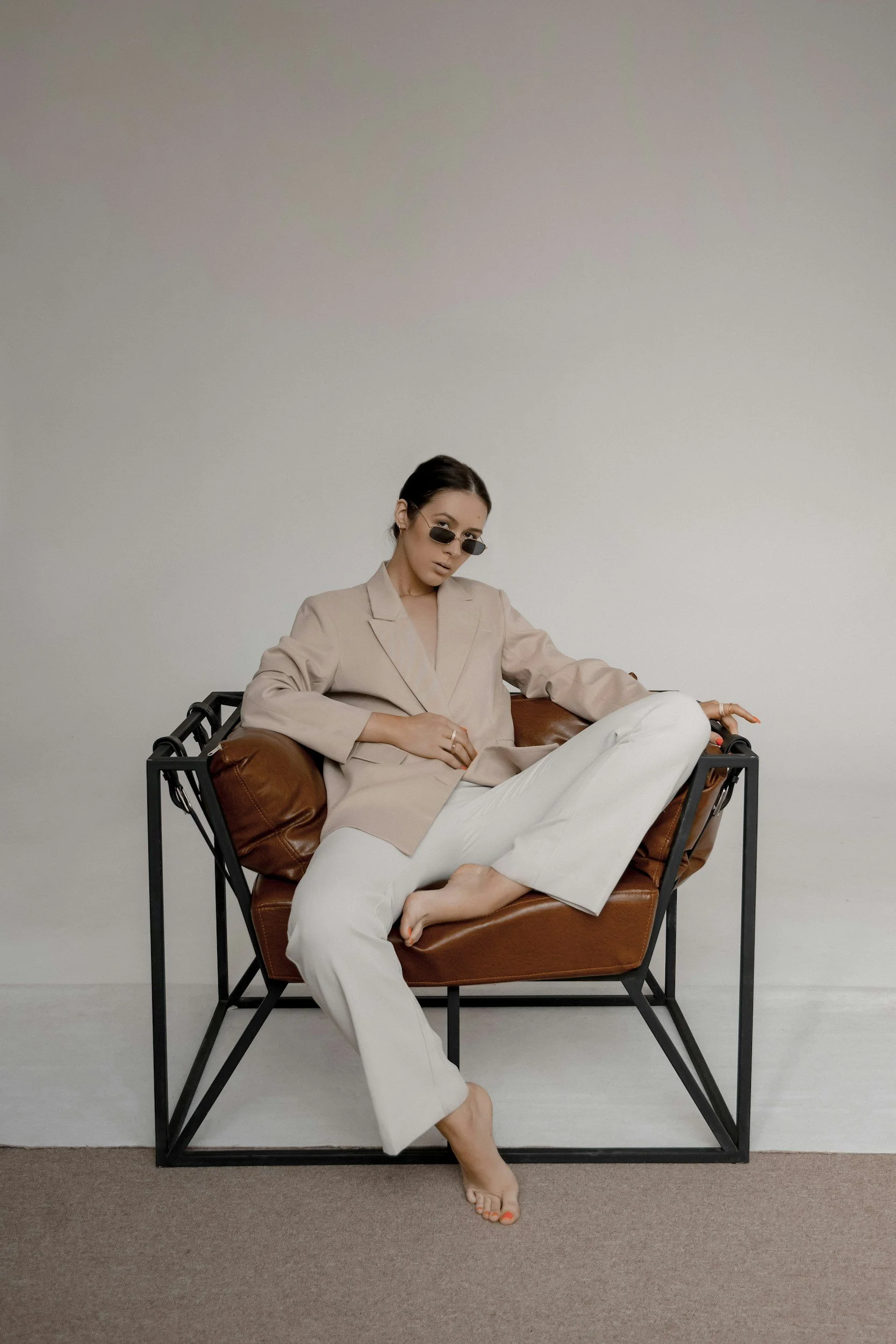A woman sitting on a modern metal and leather chair, dressed in a beige blazer and white pants, wearing sunglasses, with her legs crossed, bare feet, and side-swept dark hair, against a plain light background.