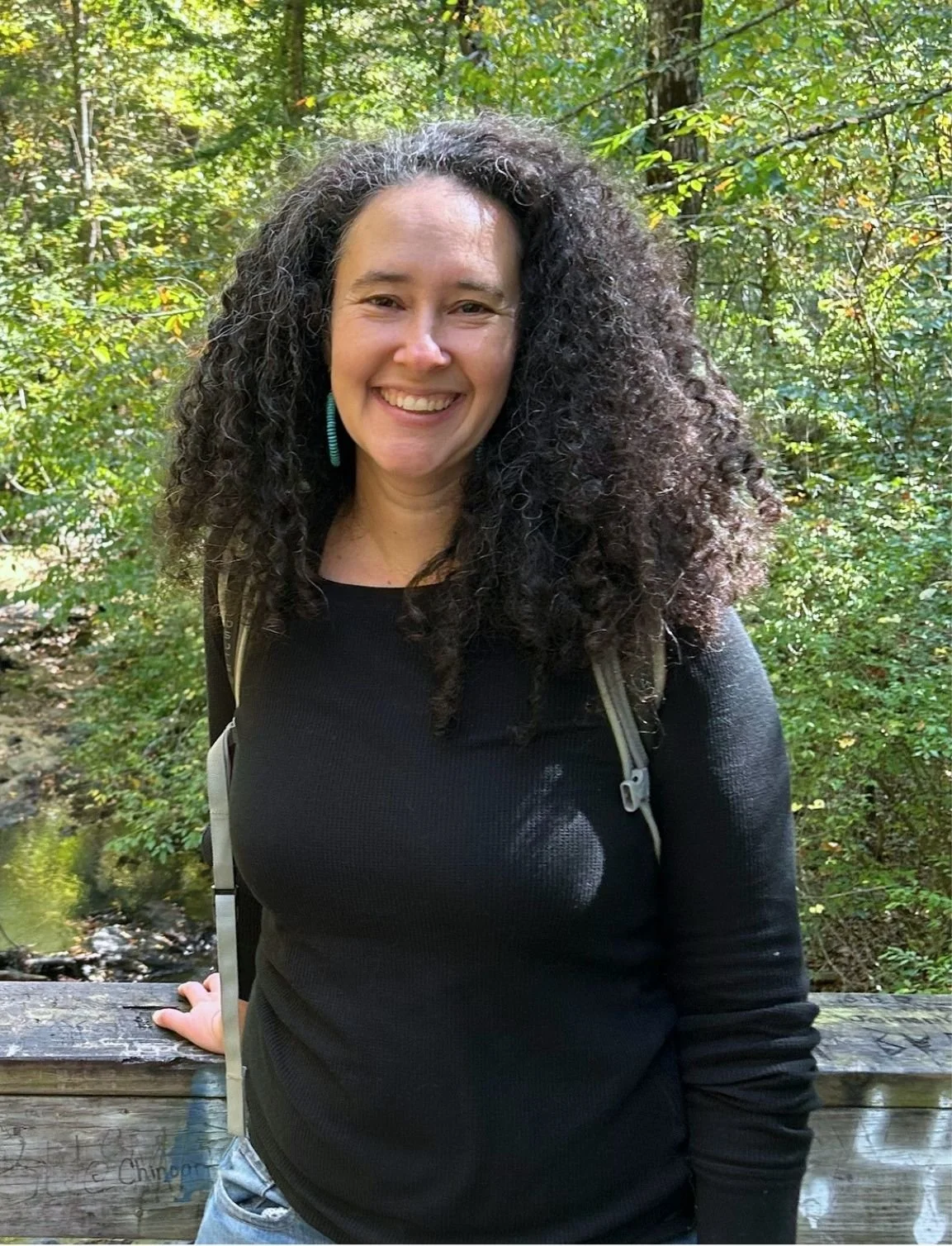 A picture of Amy Jaret, a white woman with long, curly, dark brown hair smiling outdoors in a wooded area, wearing a black long-sleeve shirt and a backpack.