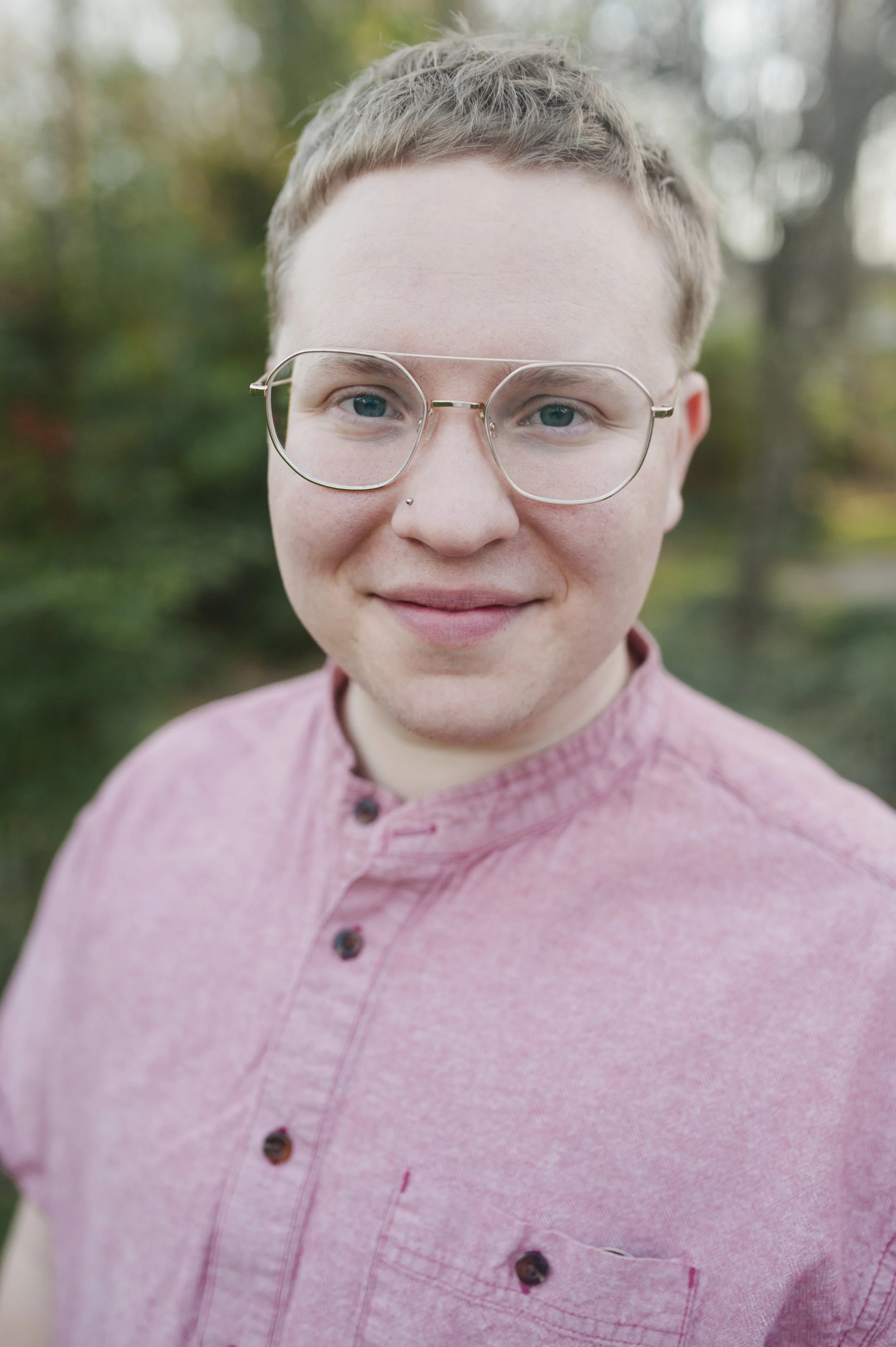 Close-up of Rabbi Eli DeWitt, a white person with light skin, blonde hair, and blue eyes, wearing large silver glasses and a pink button-up shirt, outdoors with blurred trees in the background.