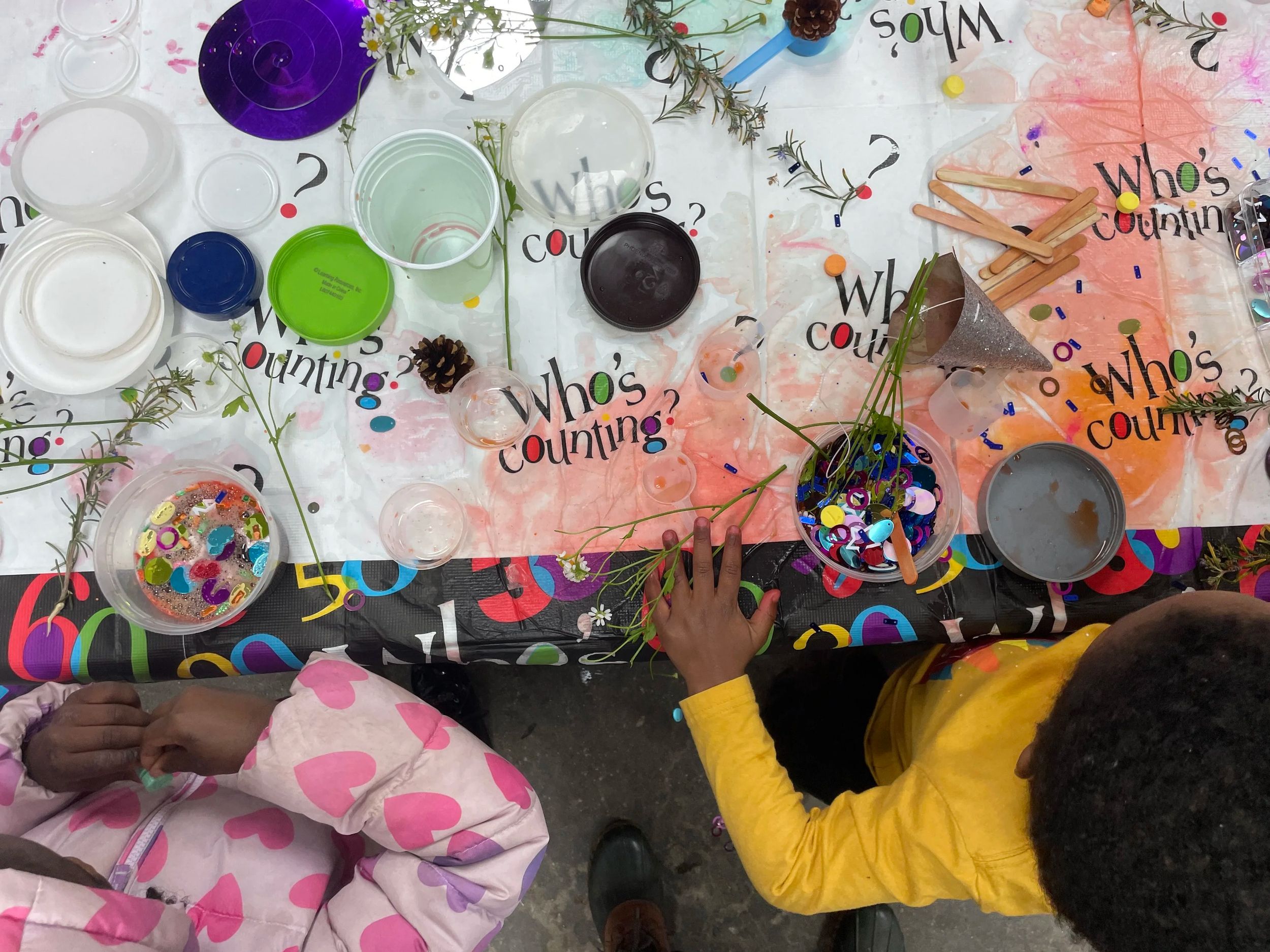 Jewish children sit at a craft table with containers of colorful confetti, sticks, pinecones, flowers, all for Tu Bishvat potion making.