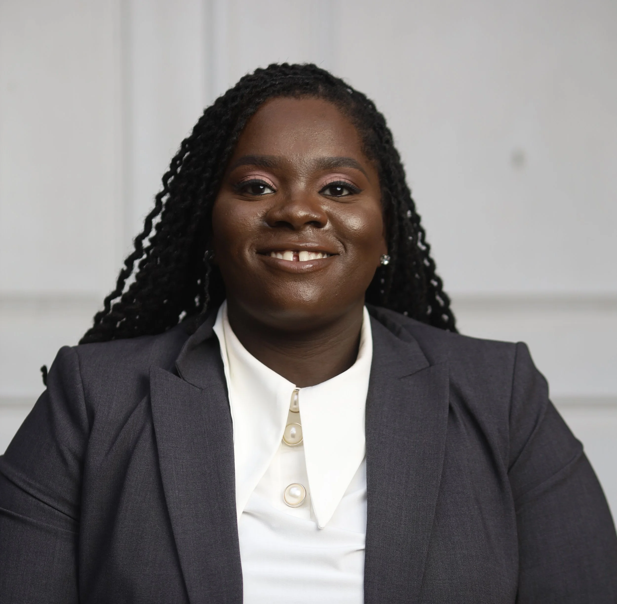 Close-up of a woman with dark skin, wearing a dark blazer, white shirt, and pearl button at the collar, smiling with visible teeth, black curly hair, and earrings, against a plain light-colored background.