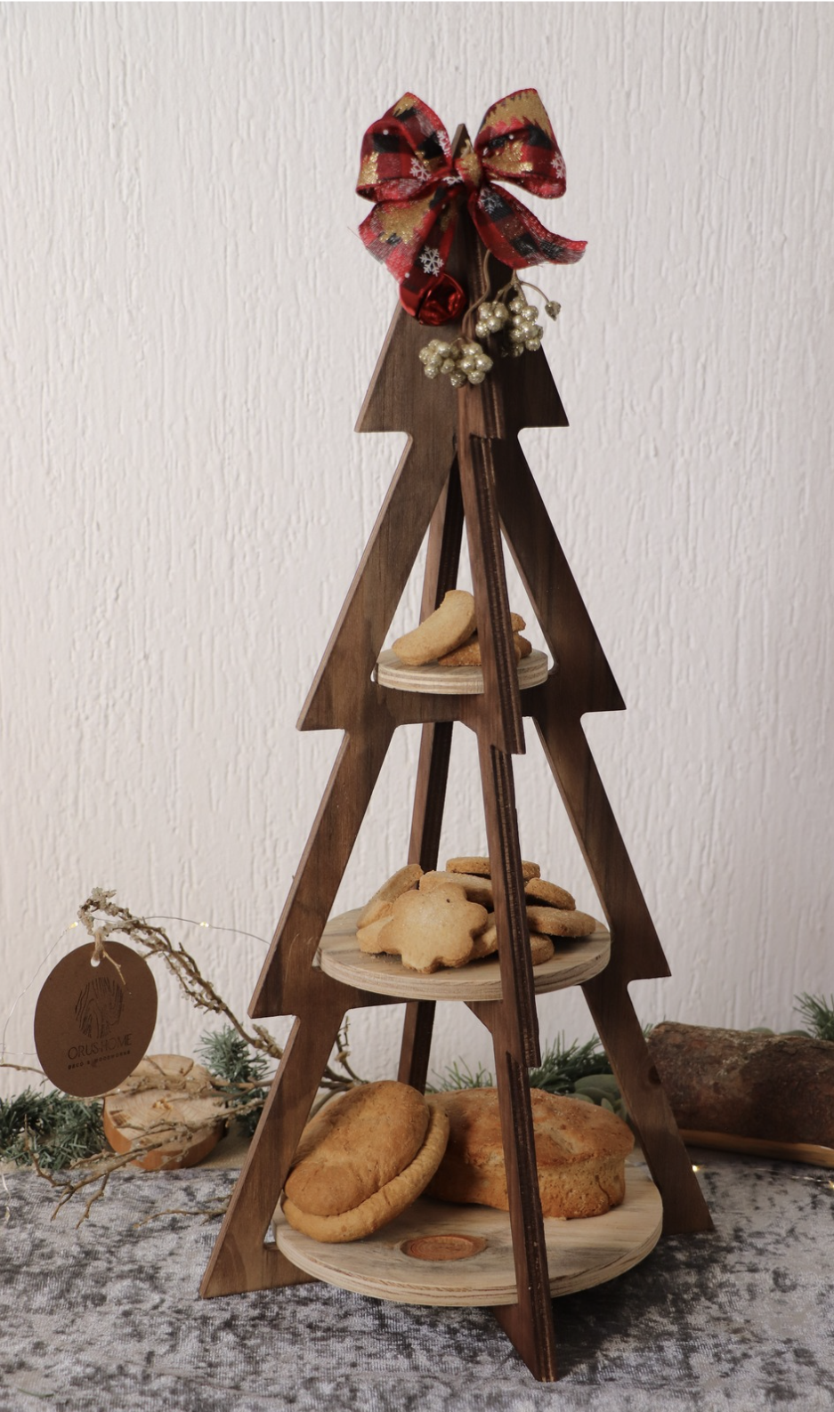 A wooden Christmas tree-shaped cookie display with three tiers of cookies, decorated with a red bow and small white ornaments, placed on a table against a white textured wall.