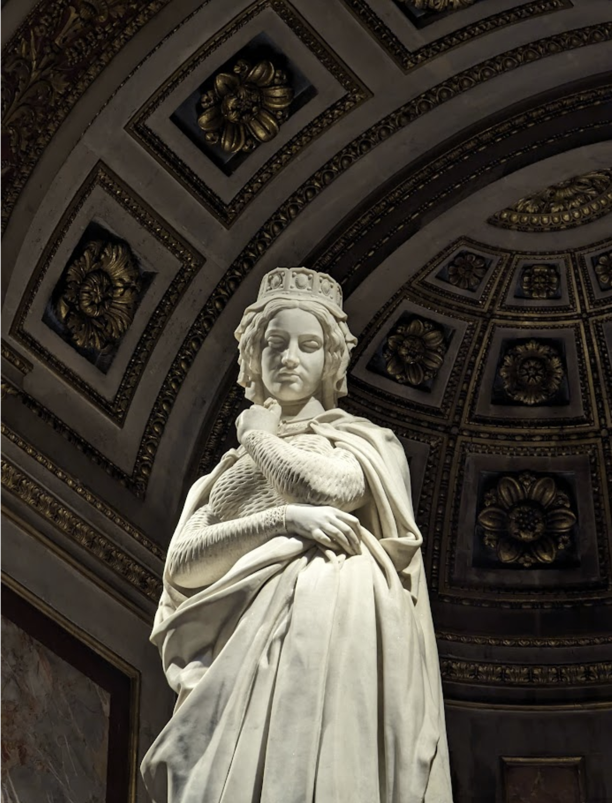 A marble statue of a woman wearing a crown, draped in clothing, standing in front of an ornate ceiling with floral decorations.