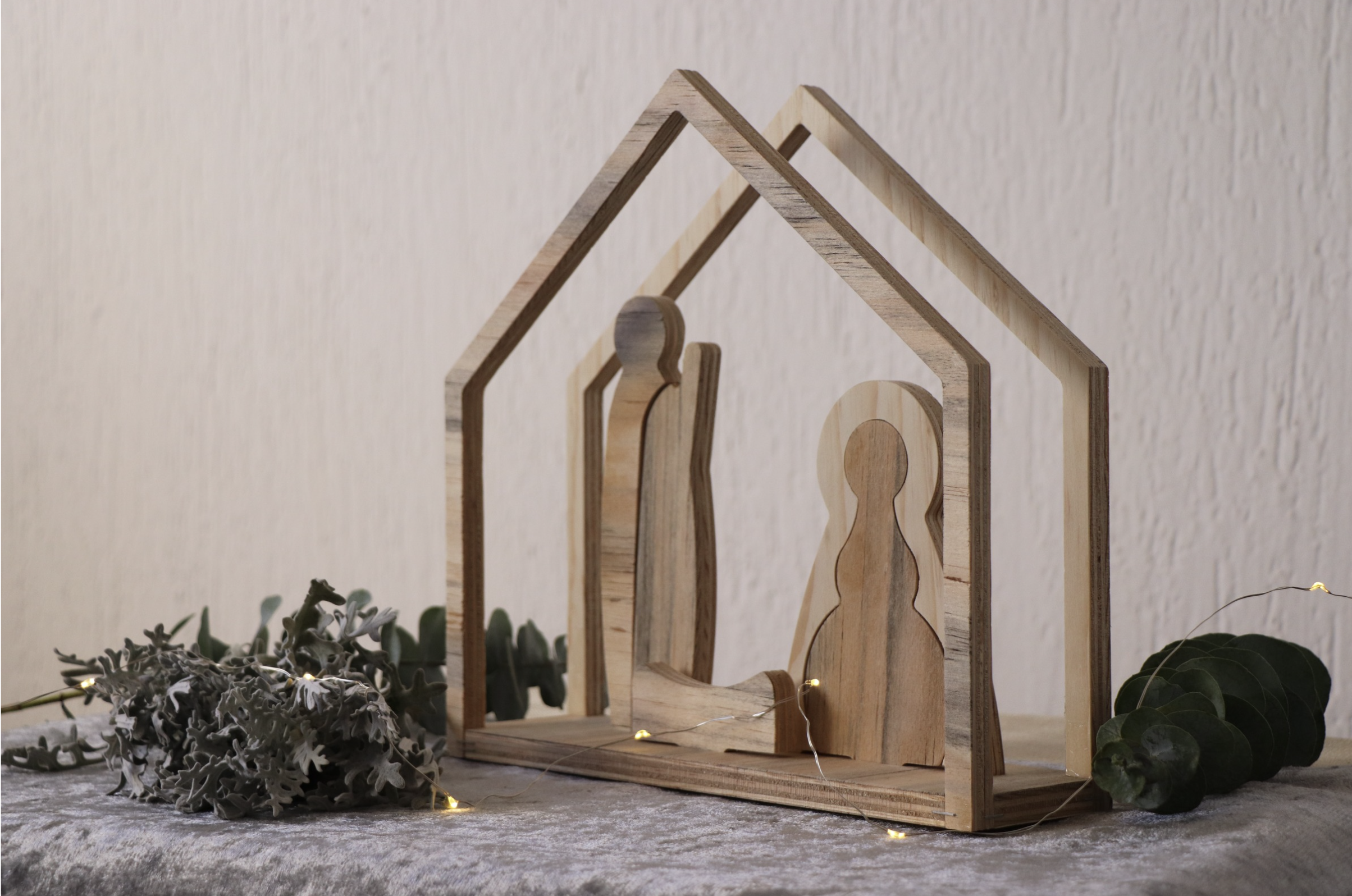 Wooden house-shaped photo display with small LED lights, surrounded by greenery and gray decorative leaves, on a light-colored textured surface.