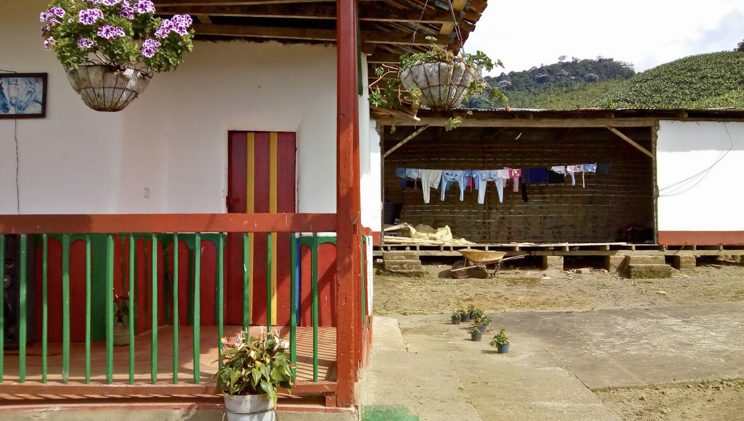 A rural outdoor scene with a colorful porch on the left side, featuring hanging flower baskets, a potted plant, and a small colorful fence. On the right side, there is a wooden structure with laundry hanging to dry, surrounded by small potted plants on the ground and a wheelbarrow. In the background, there are lush green hills under a partly cloudy sky.