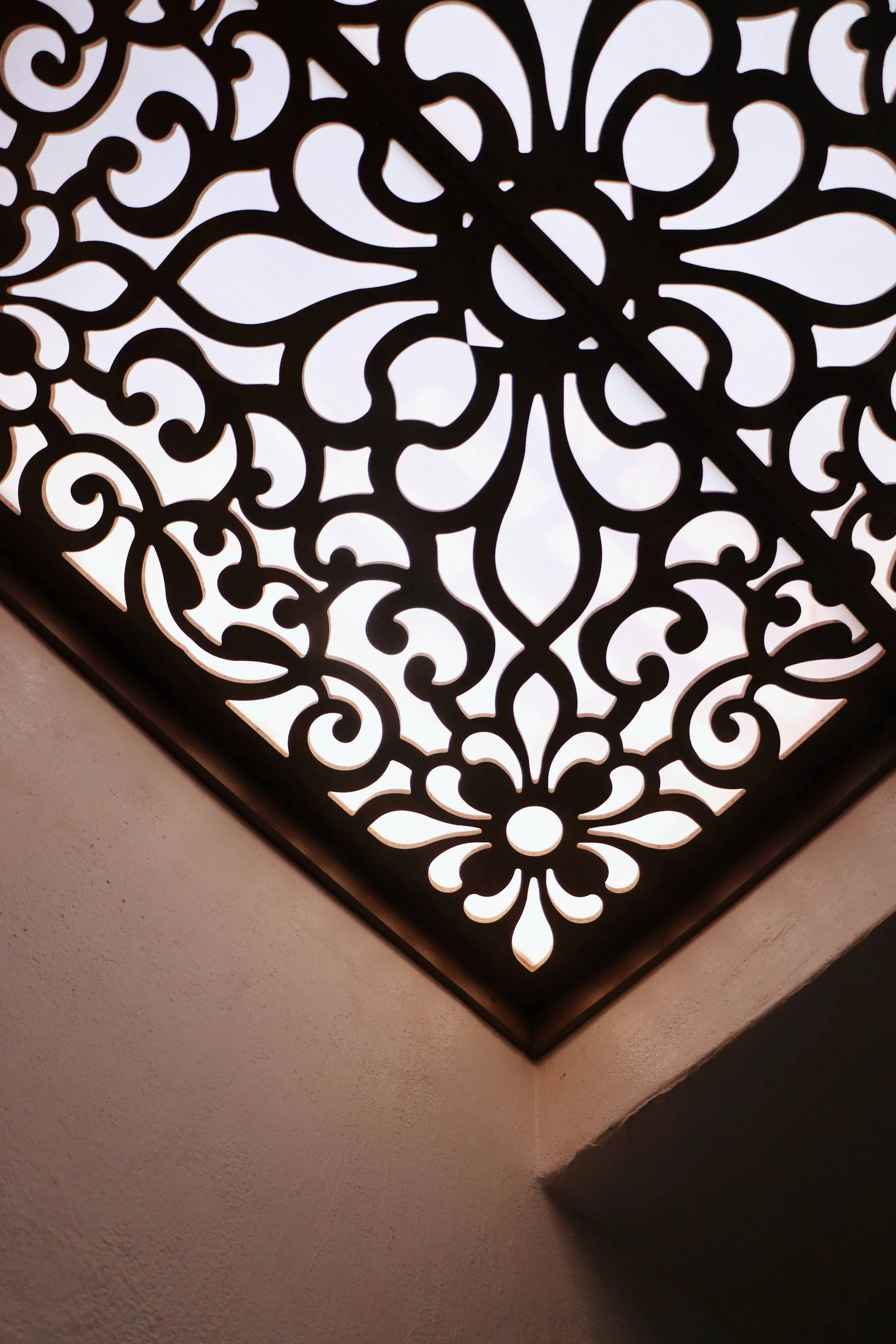 A decorative ceiling light fixture with an intricate floral and swirl pattern, with light shining through the cutouts, installed in a corner of a room with a textured beige wall.