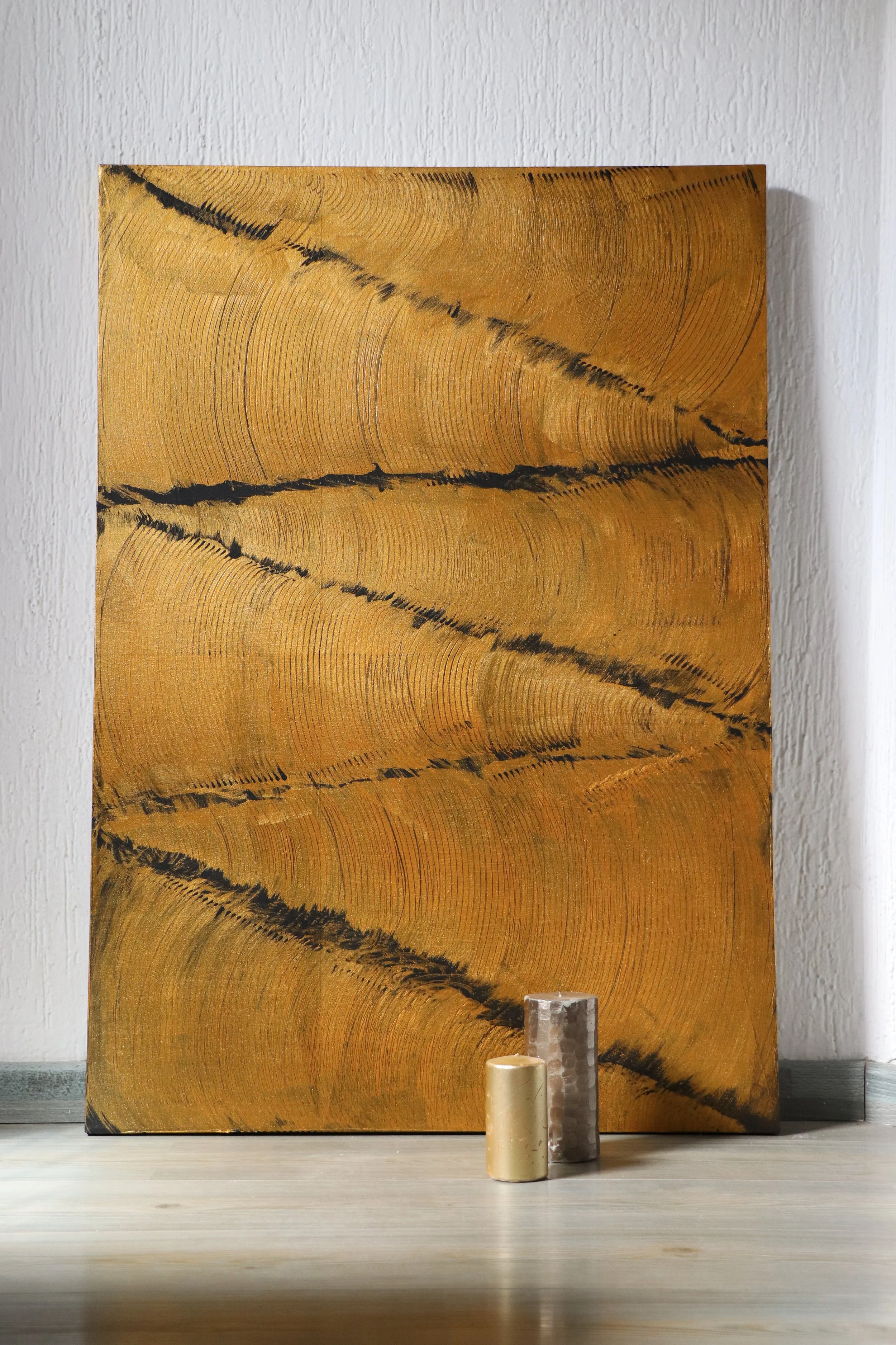 A painting of tree rings on wood leaning against a white wall, with three candles in front of it on a wooden surface.