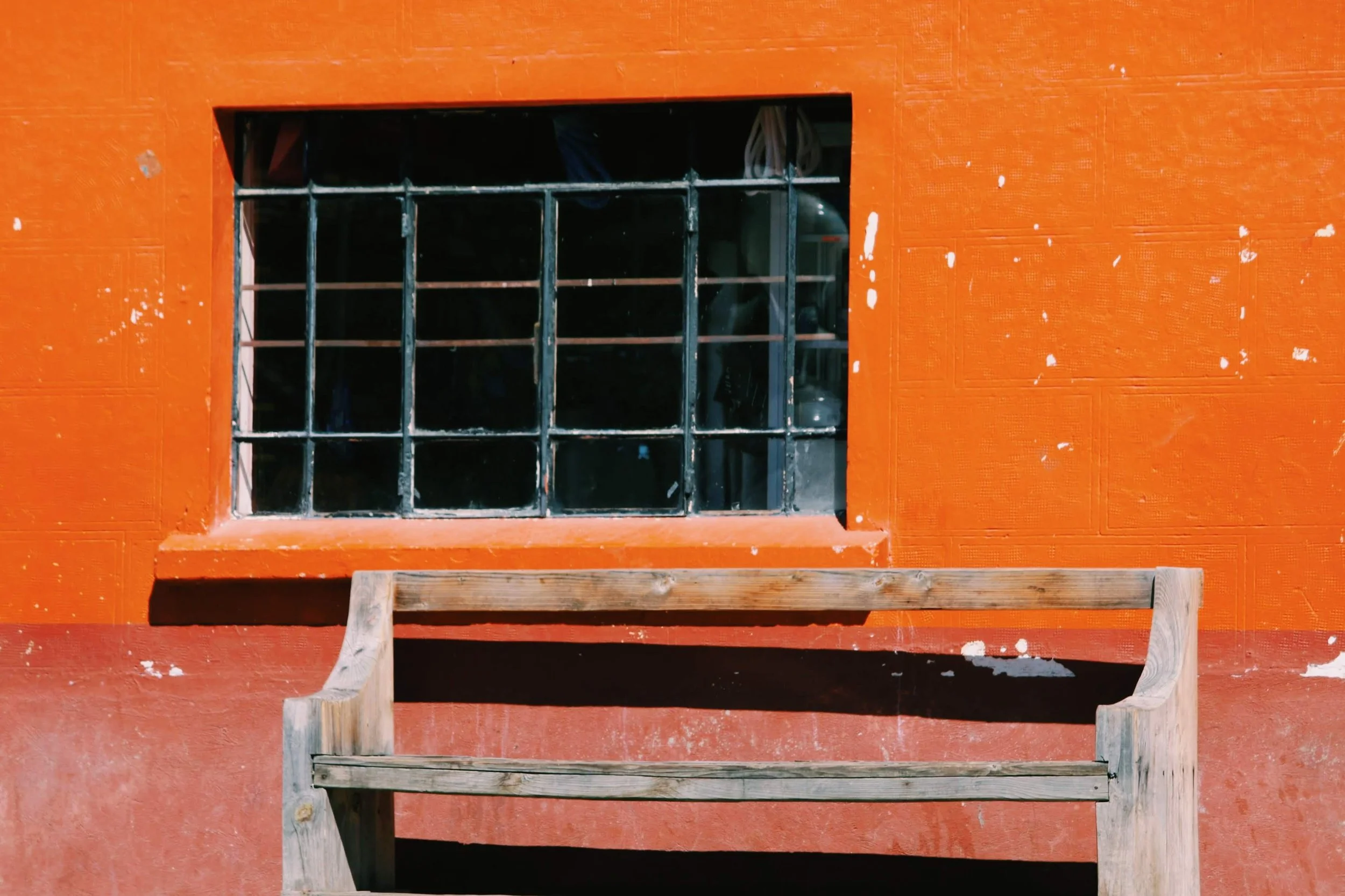 A window with black metal grill on a red textured wall, with an empty wooden bench in front of it.