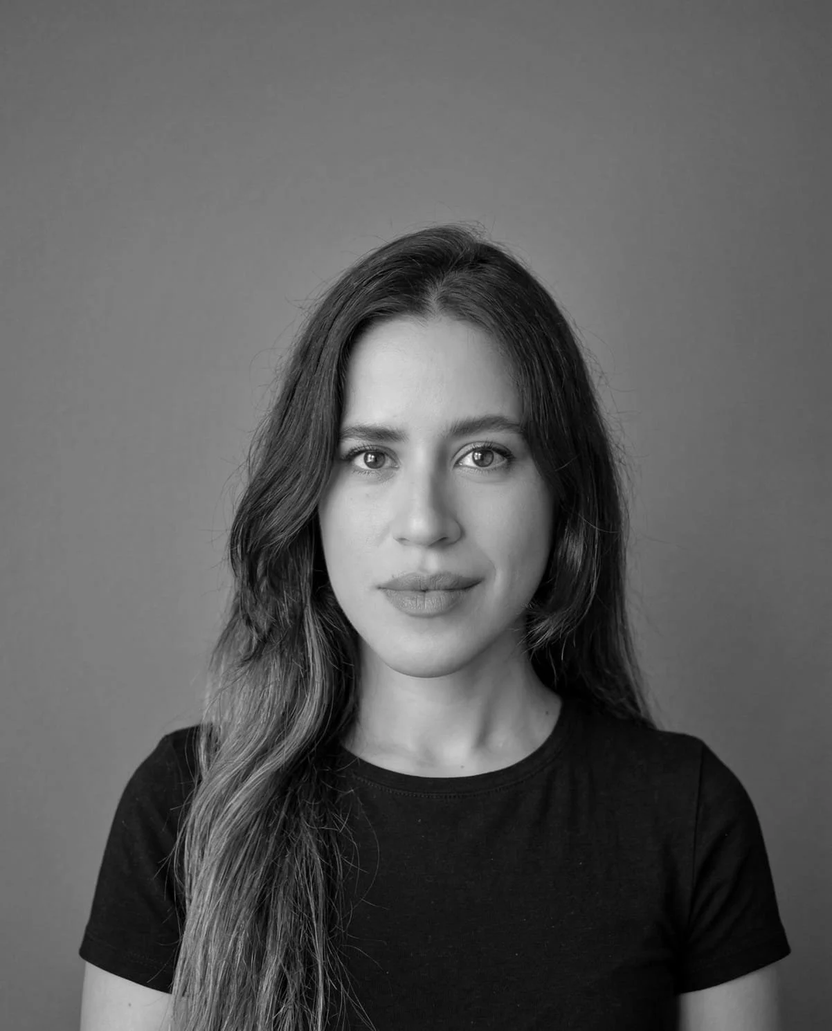 Black and white portrait of a woman with long wavy hair, looking directly at the camera, wearing a black t-shirt, against a plain background.