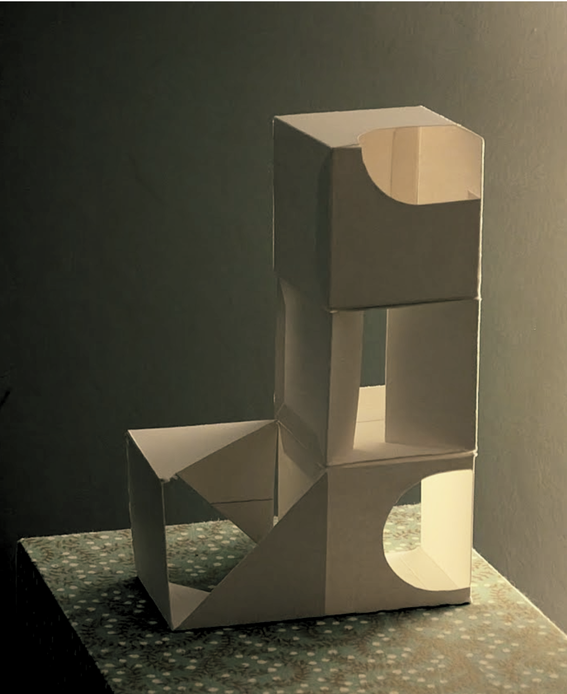 A small sculpture made of white, glossy material, resembling a stylized robot with geometric shapes, sitting on a patterned surface against a dark wall.