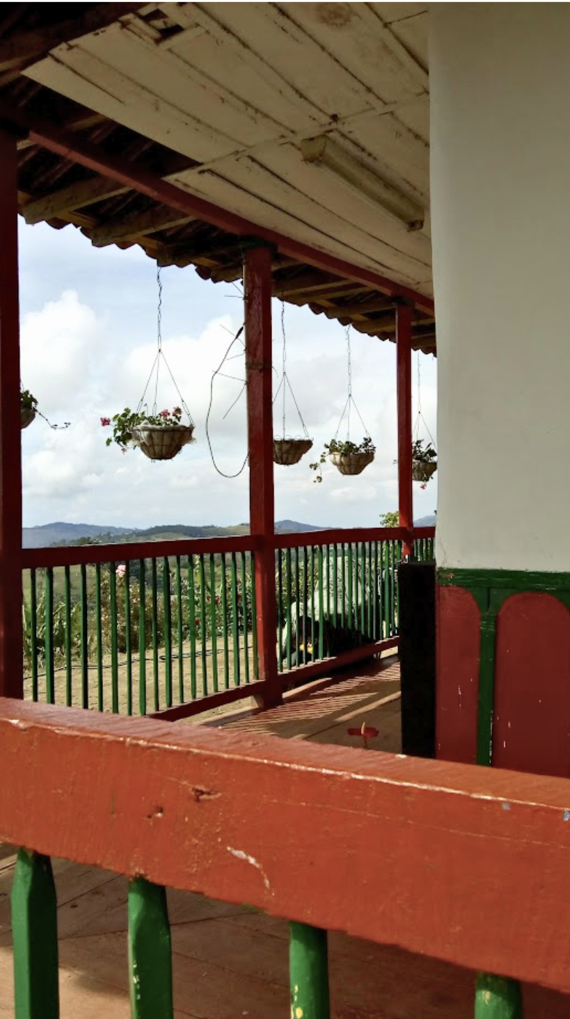View from a covered balcony with hanging flower baskets, red and green railing, overlooking a landscape with hills and cloudy sky.