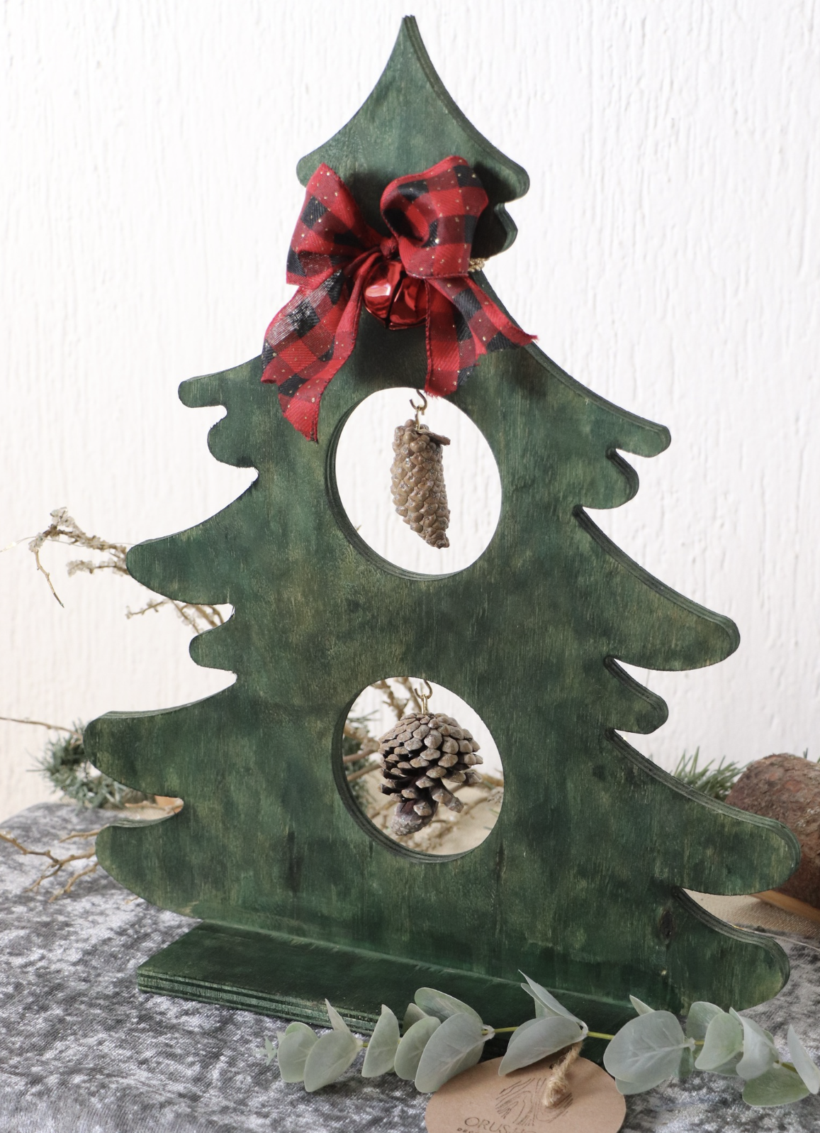 Decorative green wooden Christmas tree with cutouts, adorned with a red and black checkered ribbon bow, pine cone ornaments hanging through the cutouts, and surrounded by dried eucalyptus leaves and other natural elements.