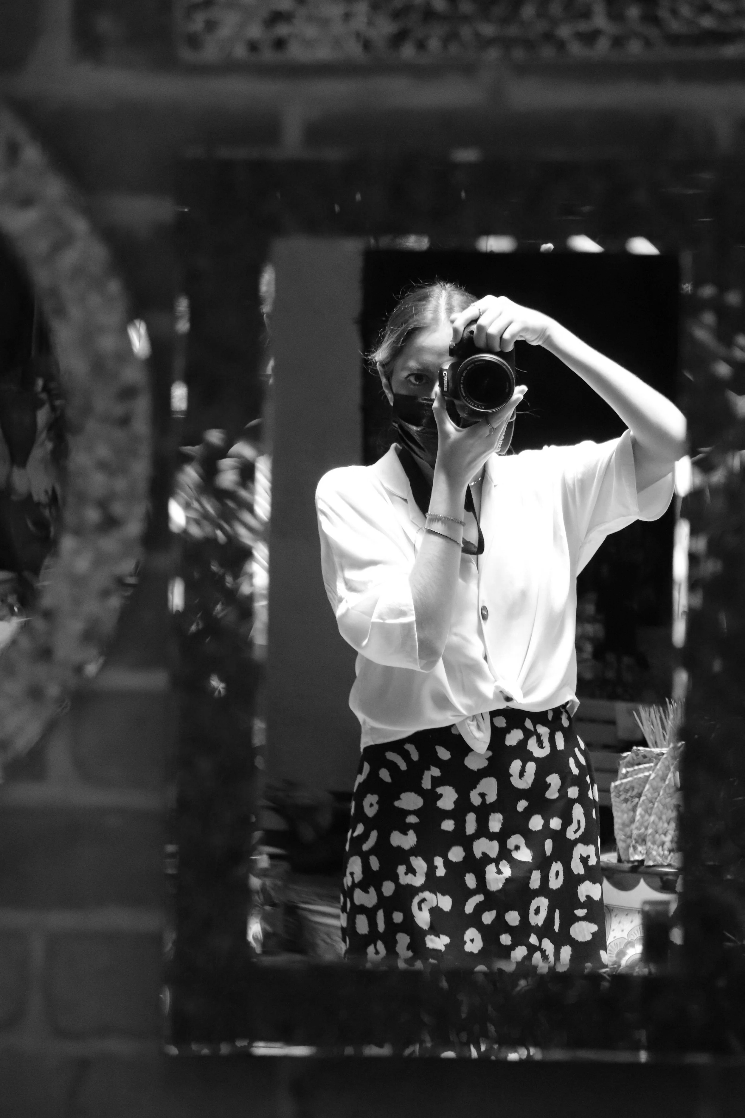A woman taking a selfie with a camera in front of a mirror, wearing a white top, animal print skirt, and a face mask, in a setting that appears to be a store or market.