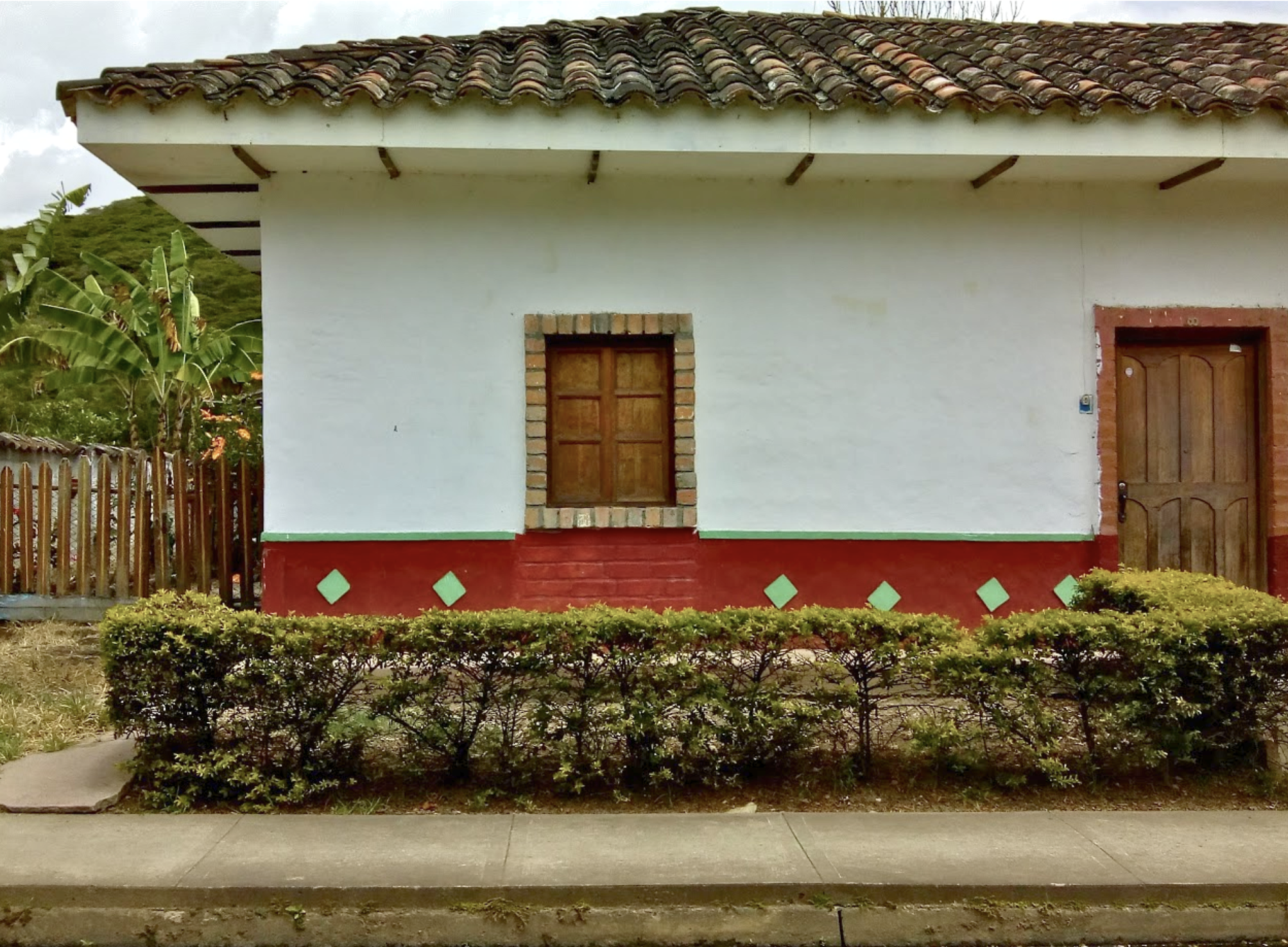 A white house with a red base, brick window frame, and wooden door. The house has a tiled roof and a small front yard with bushes and a sidewalk.