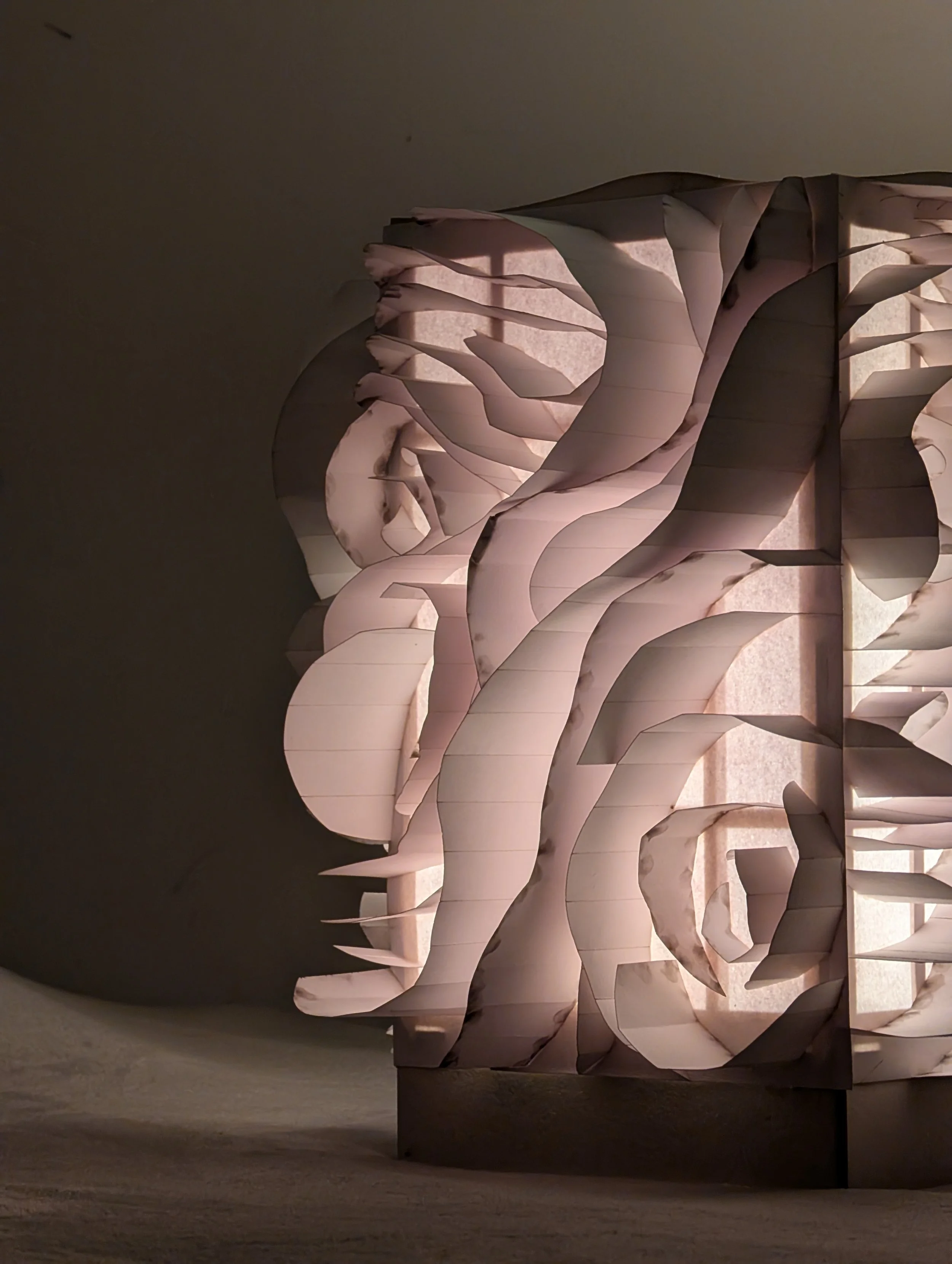 A white sculptural installation resembling a flowing, layered design, possibly made of paper or fabric, set against a dark background with lighting enhancing its contours.