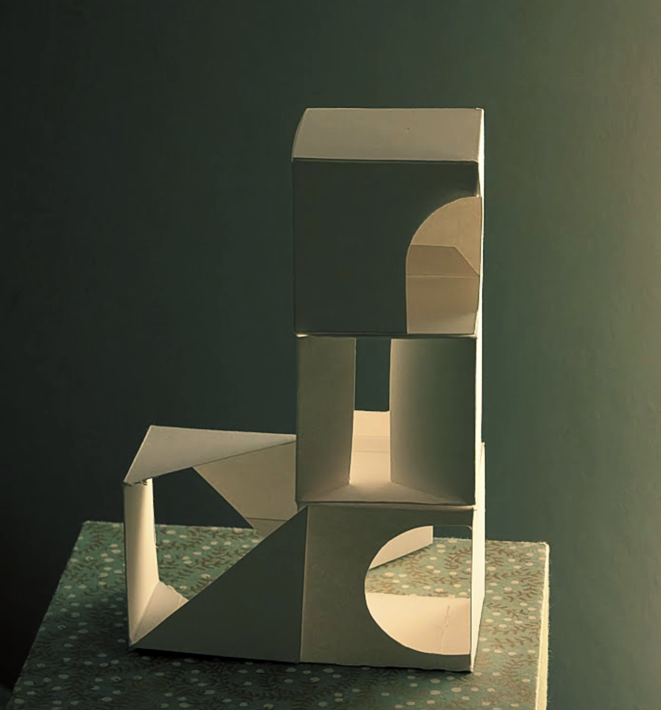 An abstract paper sculpture spelling out the word 'LOVE' with the 'L' and 'V' lying flat and the 'O' and 'E' standing upright, placed on a patterned surface against dark background.
