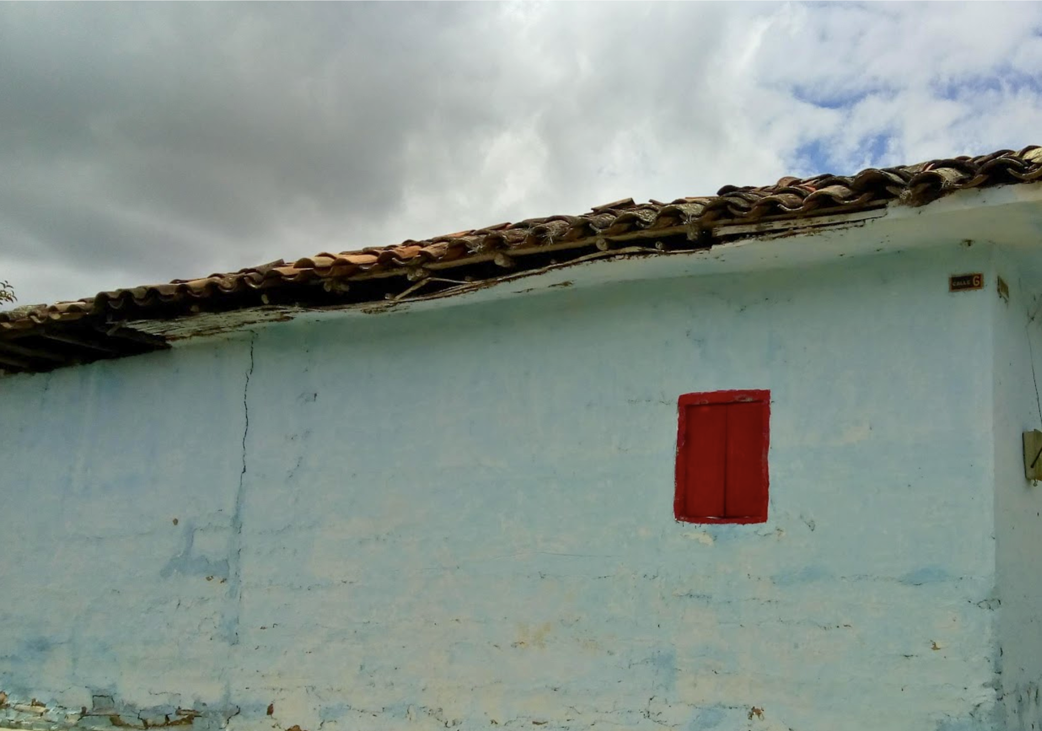 A white wall with cracks and peeling paint, a red door, and a small rectangular sign with the number 6. The wall has a rough texture and some dirt near the bottom. The roof has traditional clay tiles, some of which are damaged.
