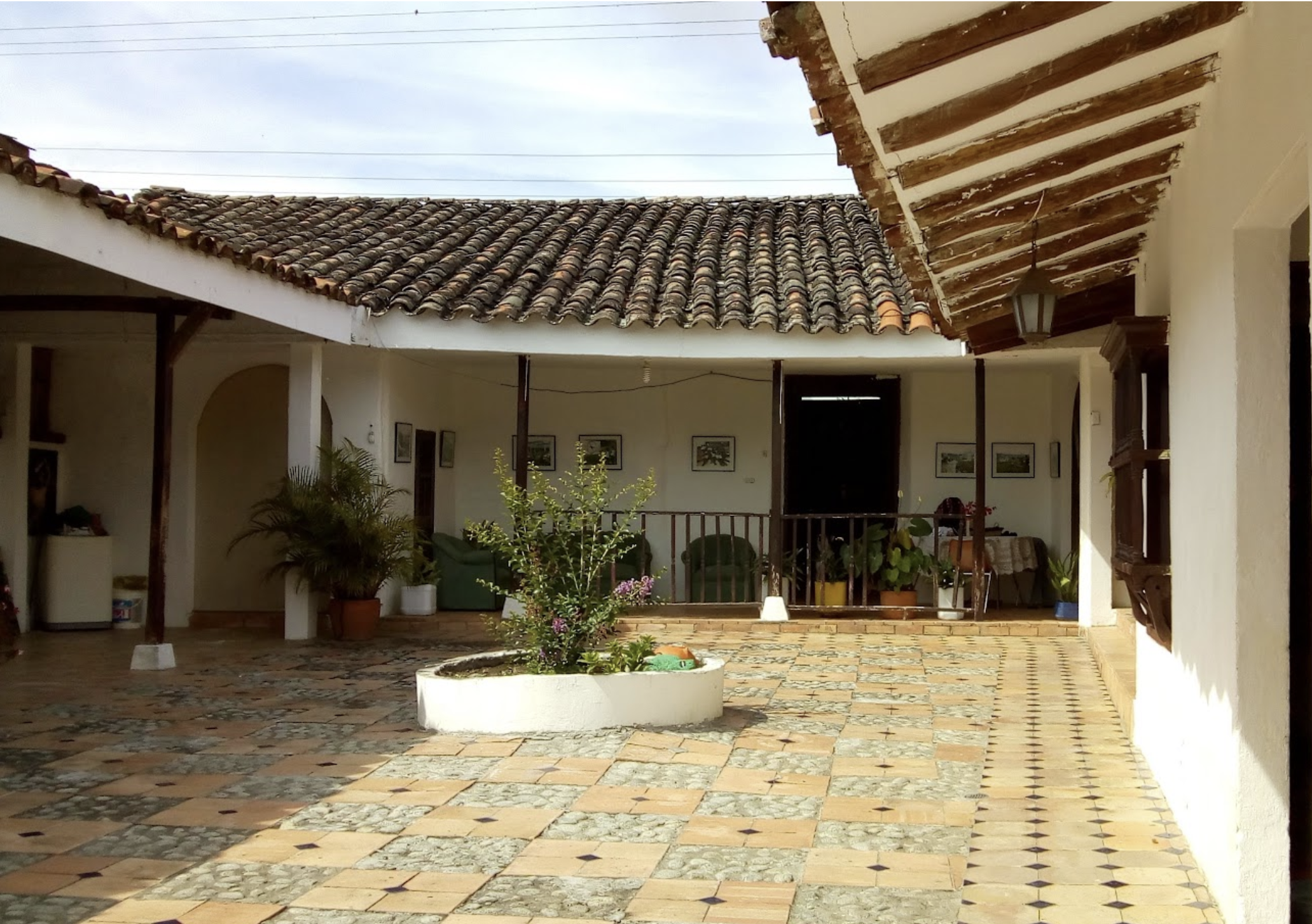A covered patio area with a tiled roof, potted plants, and framed artwork on the wall. The floor is made of patterned tiles, and the patio is open-air with a view of the sky and electrical wires.