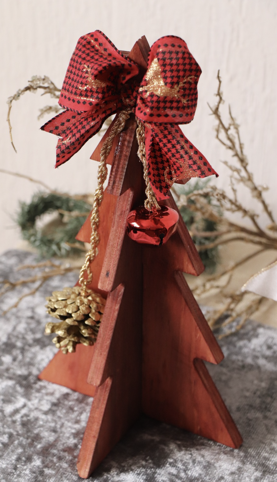 Decorative wooden Christmas tree ornament adorned with a red and black plaid bow, gold tassel, gold pinecone, and shiny red bell.