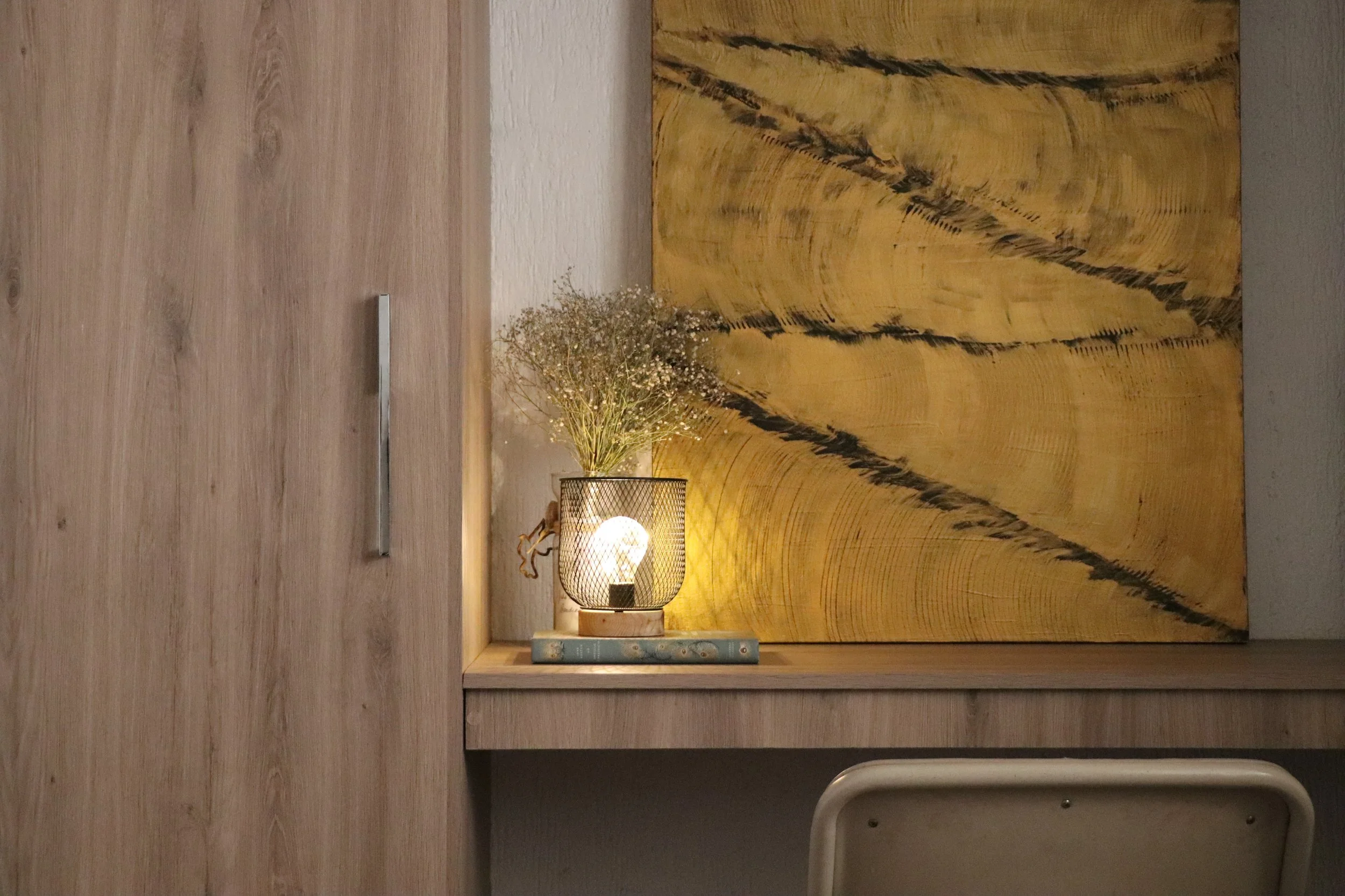A decorative indoor shelf with a wooden background, featuring a table lamp with a woven shade, a small dried plant, a closed book, and a large abstract artwork with yellow and black streaks on the wall.