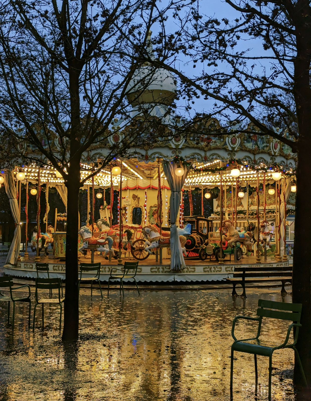 Faintly lit vintage carousel with horses and carriages on a rainy evening, partially obscured by leafless trees.